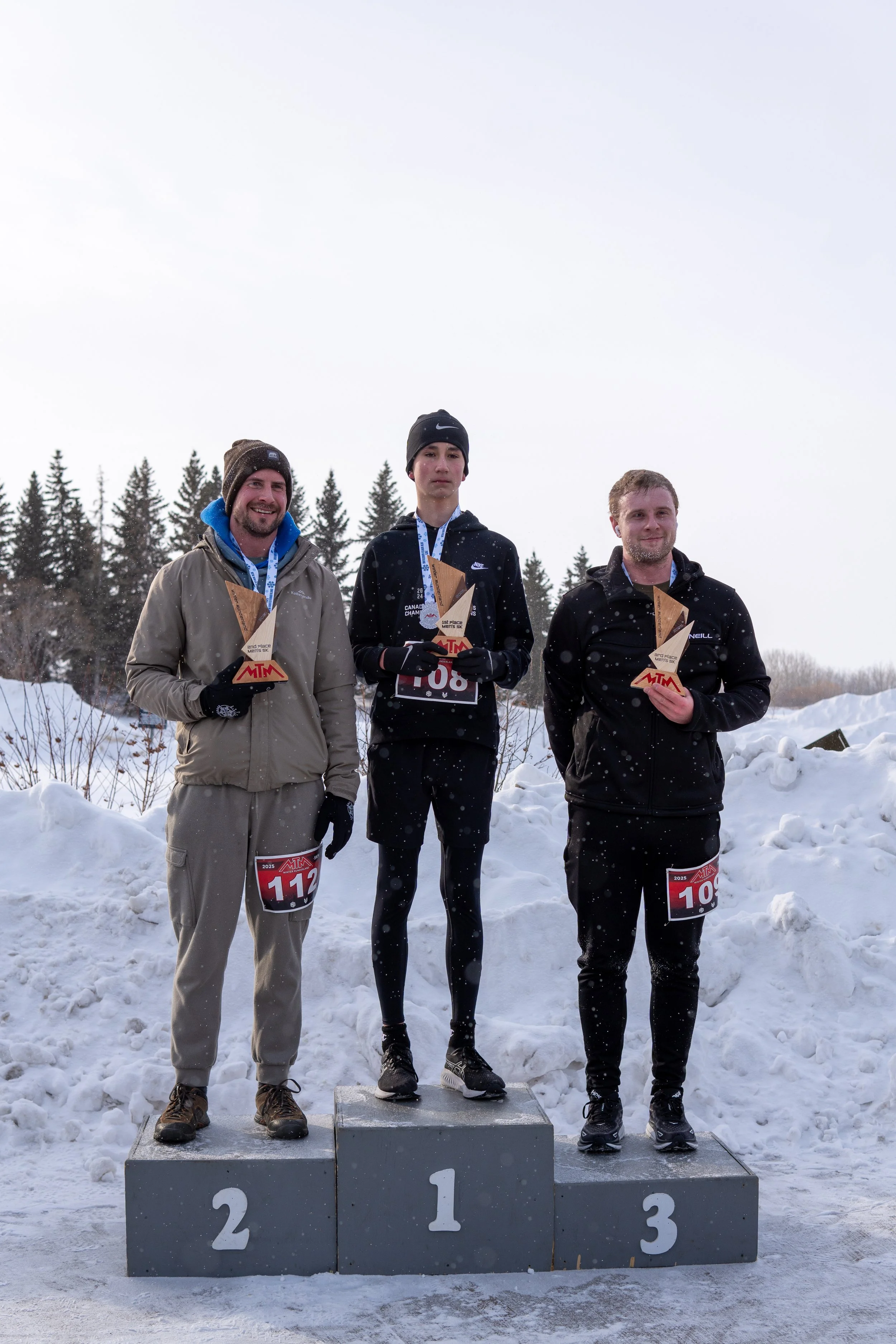 Three male athletes standing on a winners' podium outdoors in snowy weather, holding trophies, with a background of snow and trees, celebrating their finishing positions in a winter race.