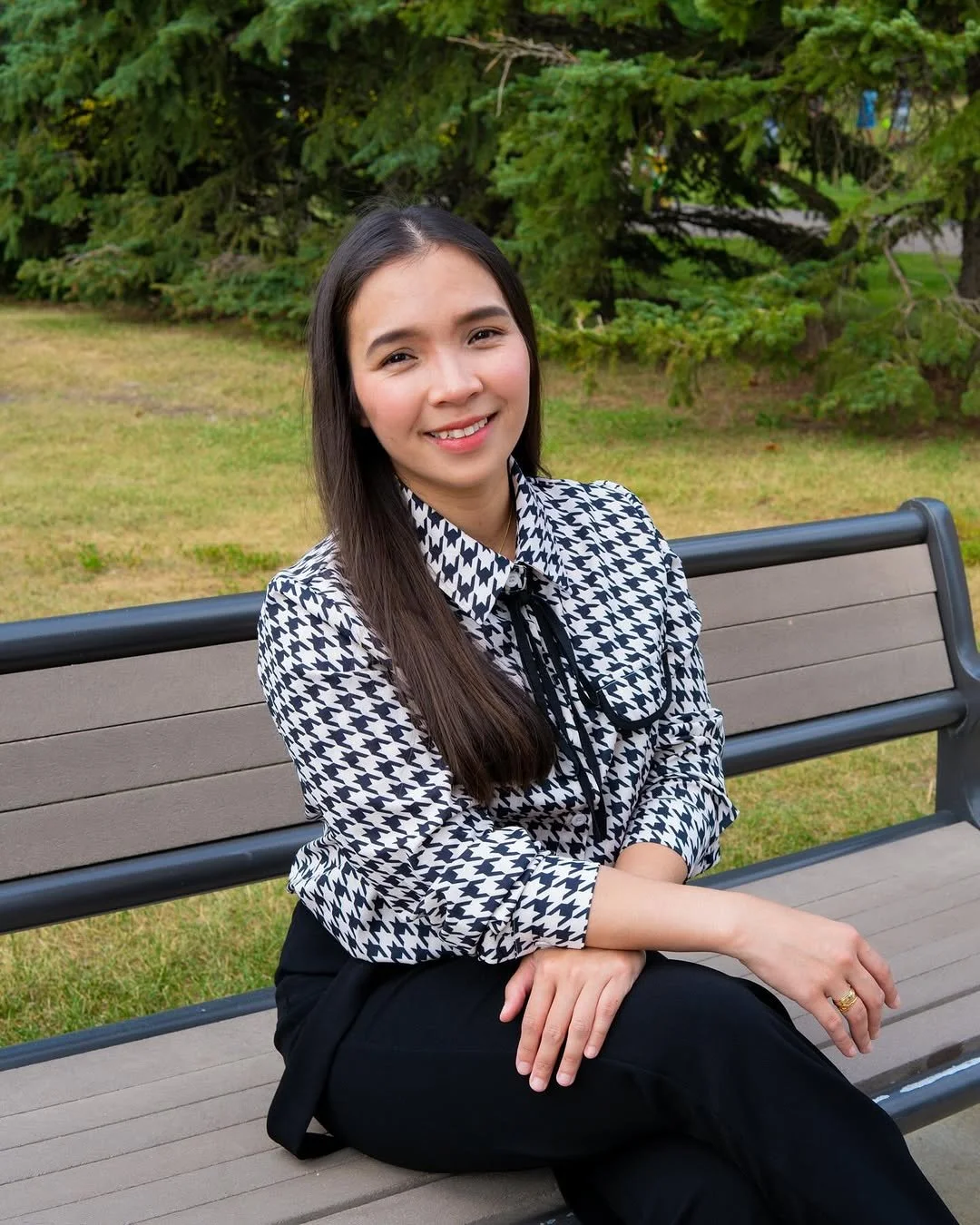 A woman with long dark hair, smiling and wearing a black and white houndstooth blouse with a black bow tie, sits on a park bench in front of green trees and grass.