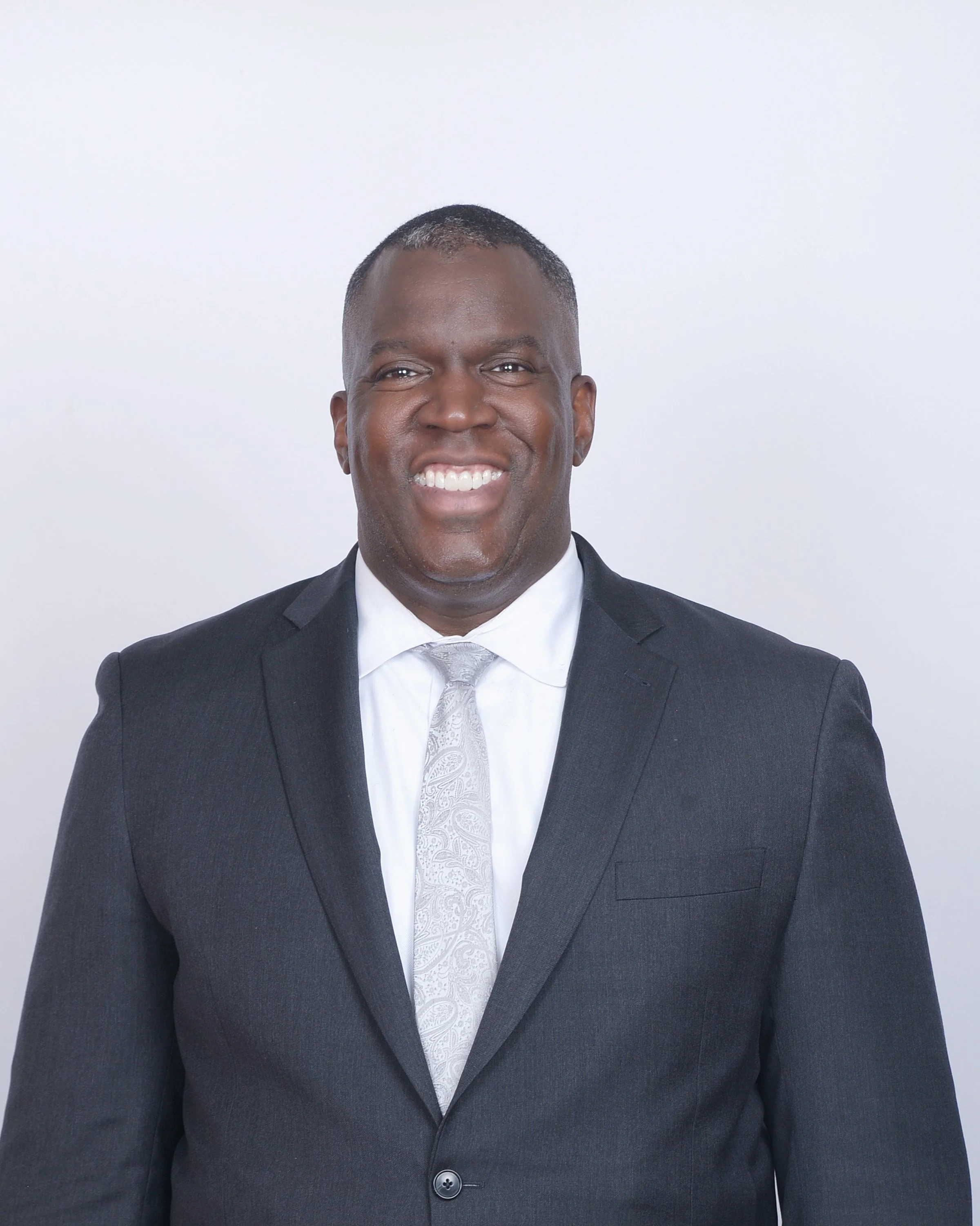 Professional man in black suit and white shirt, smiling against a plain white background.