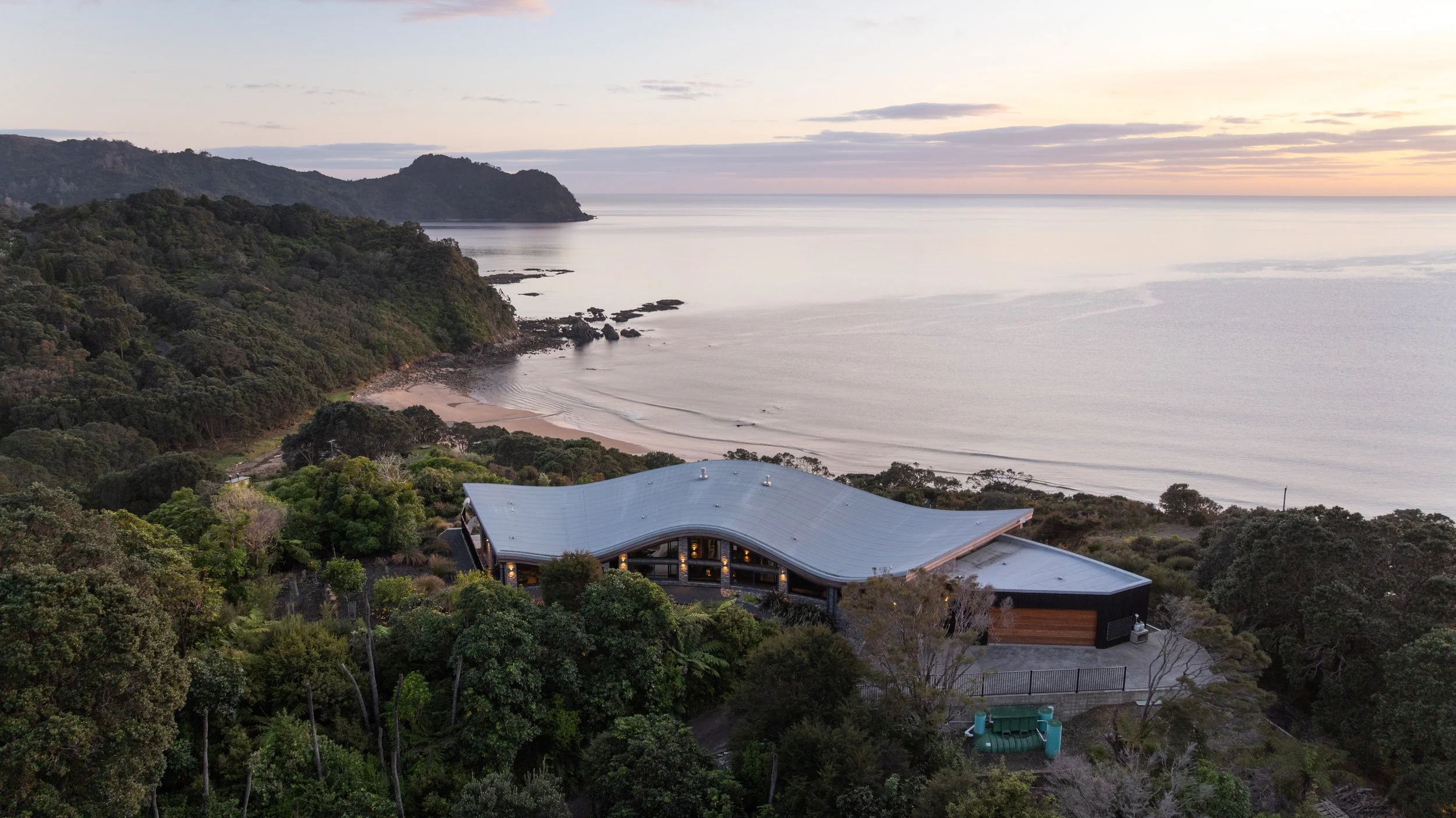 A modern house with a curved roof located on a hillside overlooking the ocean at sunset, surrounded by trees and lush greenery.