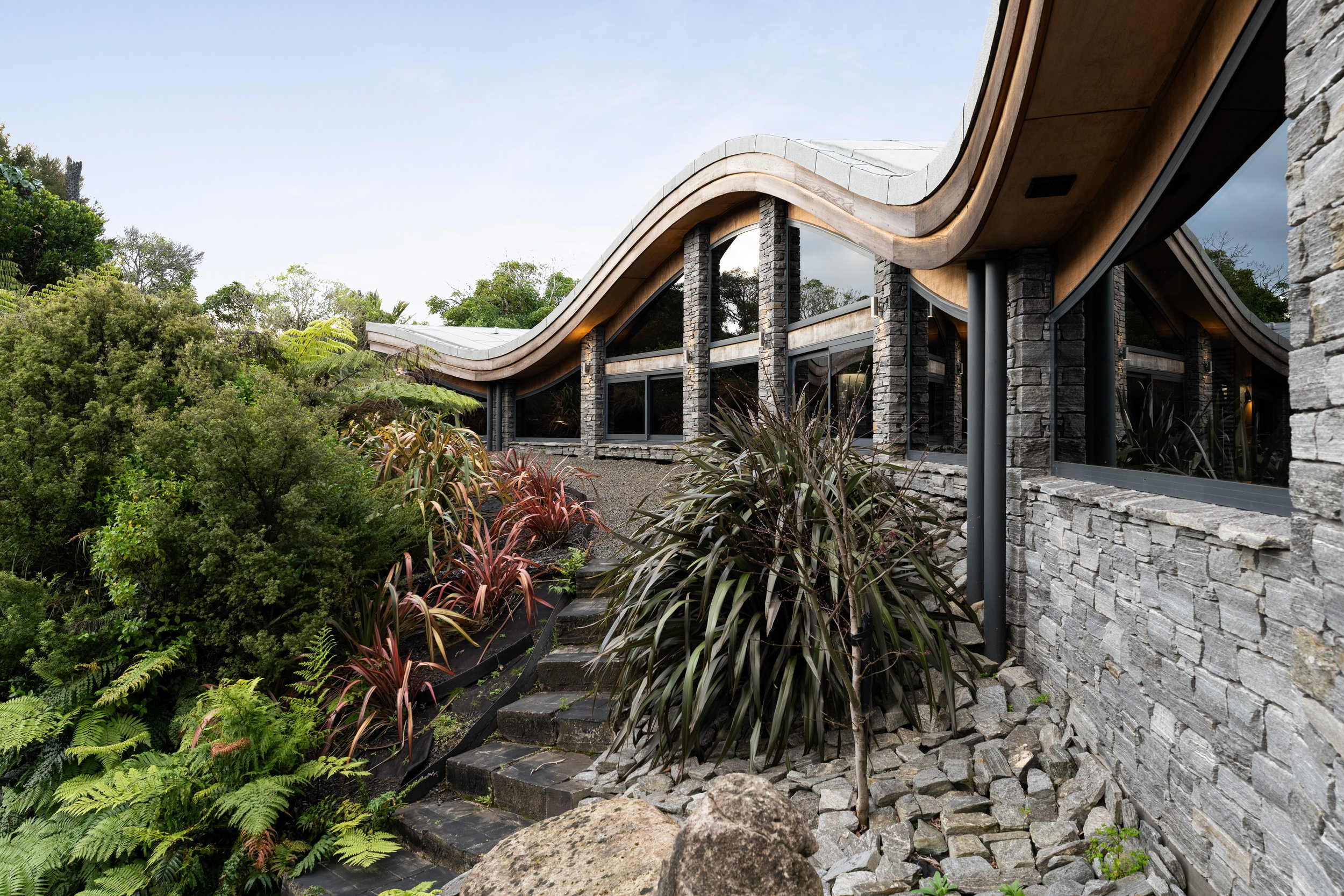 Modern house with a wavy wooden roof, stone exterior walls, large windows, surrounded by lush green plants and stairs leading up to the entrance.