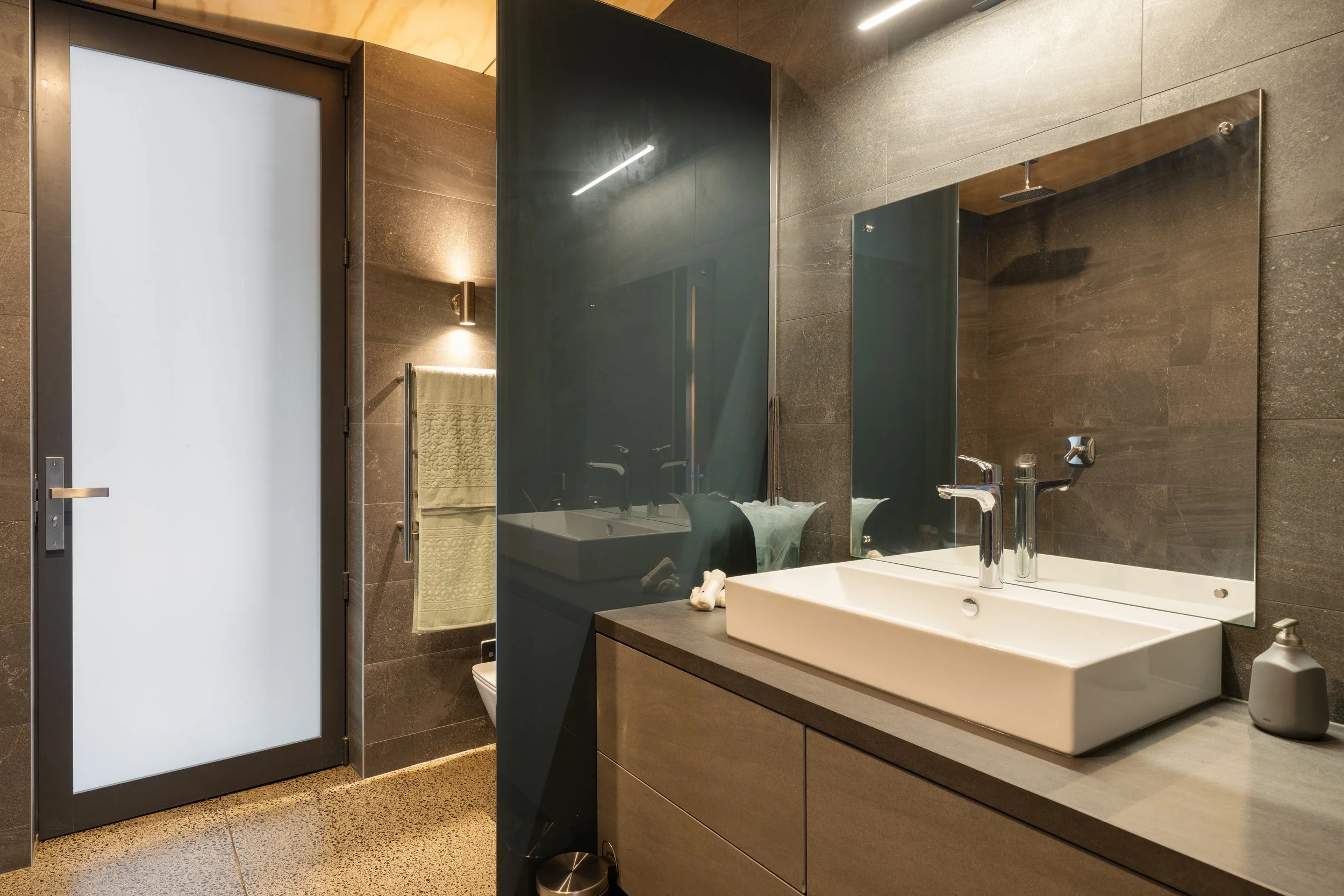 Modern bathroom with beige vanity, white rectangular sink, chrome faucet, large mirror, frosted glass shower door, brown tiled walls, and a towel on a wall-mounted rack.