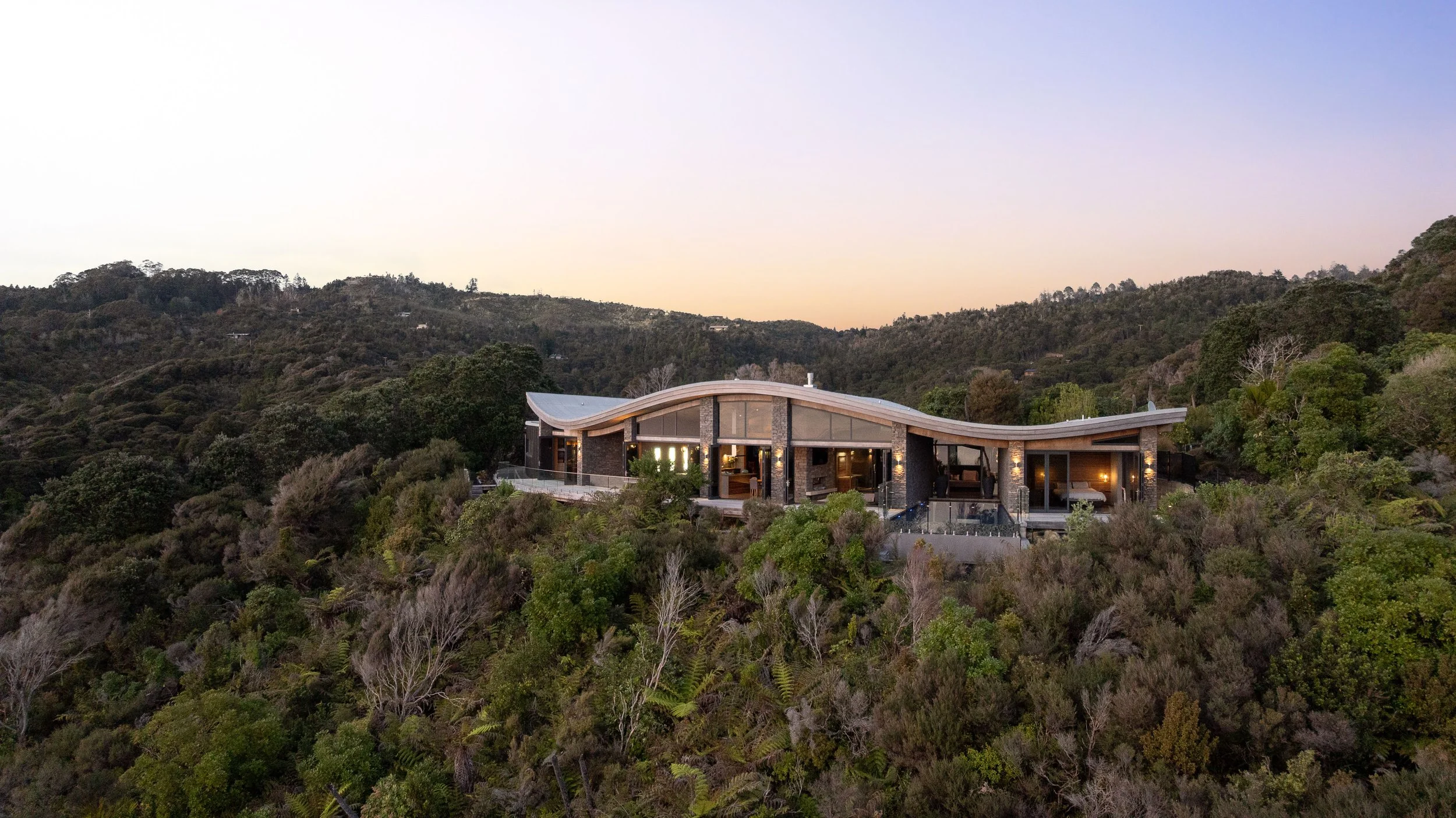 Modern house with curved roof, large windows, and outdoor patio set in a lush green hillside landscape during dusk.