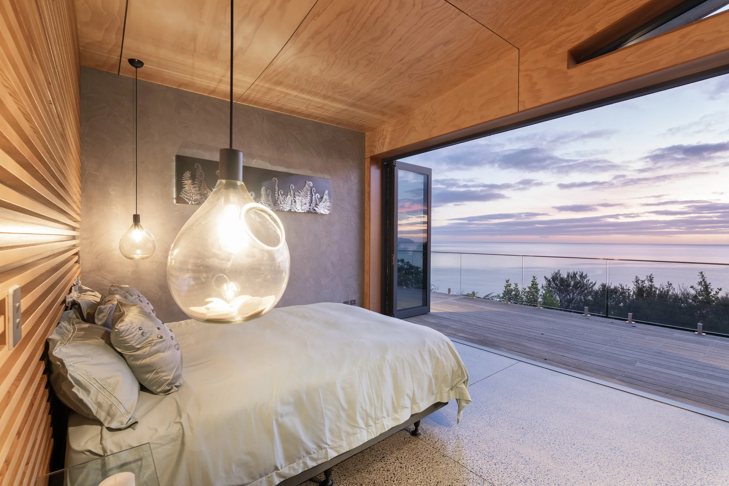 Modern bedroom with a large bed and pendant lights, open sliding glass door reveals a balcony with a view of the ocean and cloudy sky at sunrise or sunset.