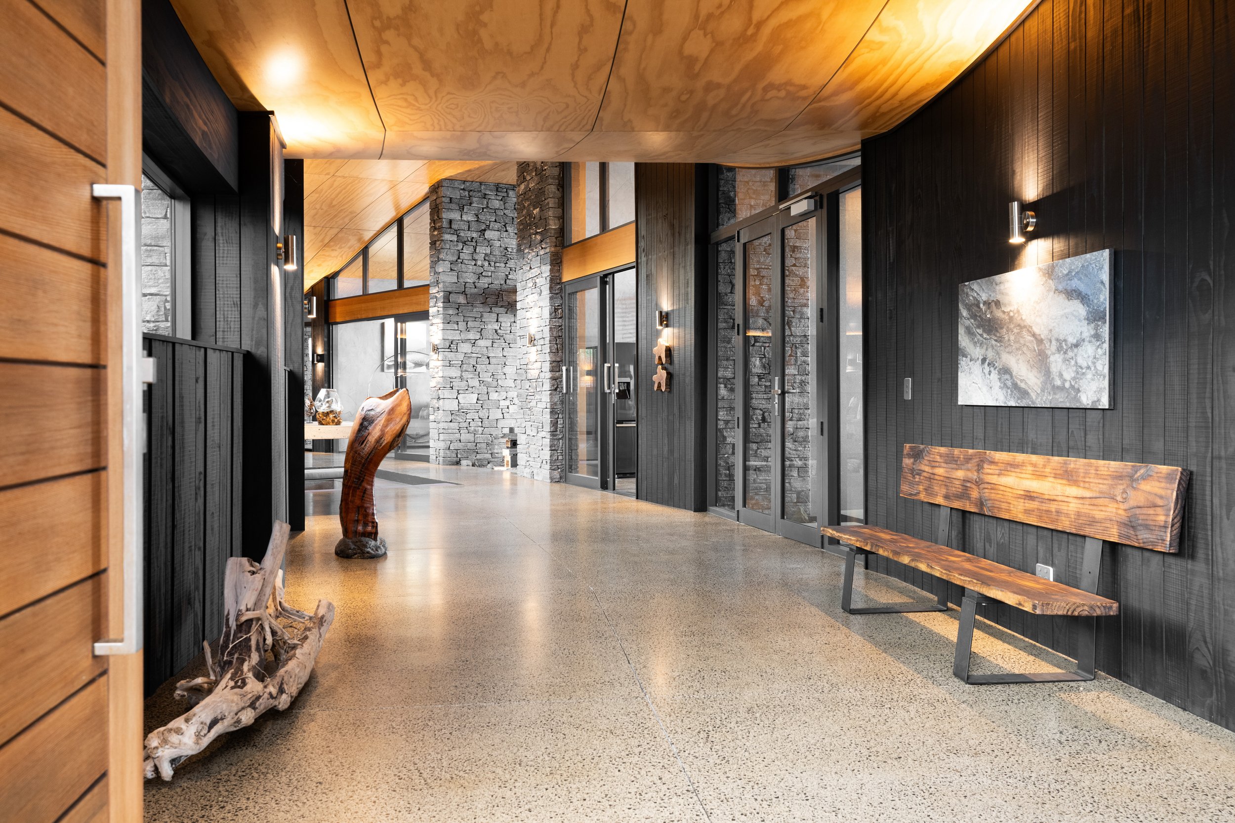 Modern indoor hallway with wood-paneled ceiling, stone and black wooden walls, and polished concrete floor. Features includes wooden and stone sculptures, a bench, and wall-mounted lights.