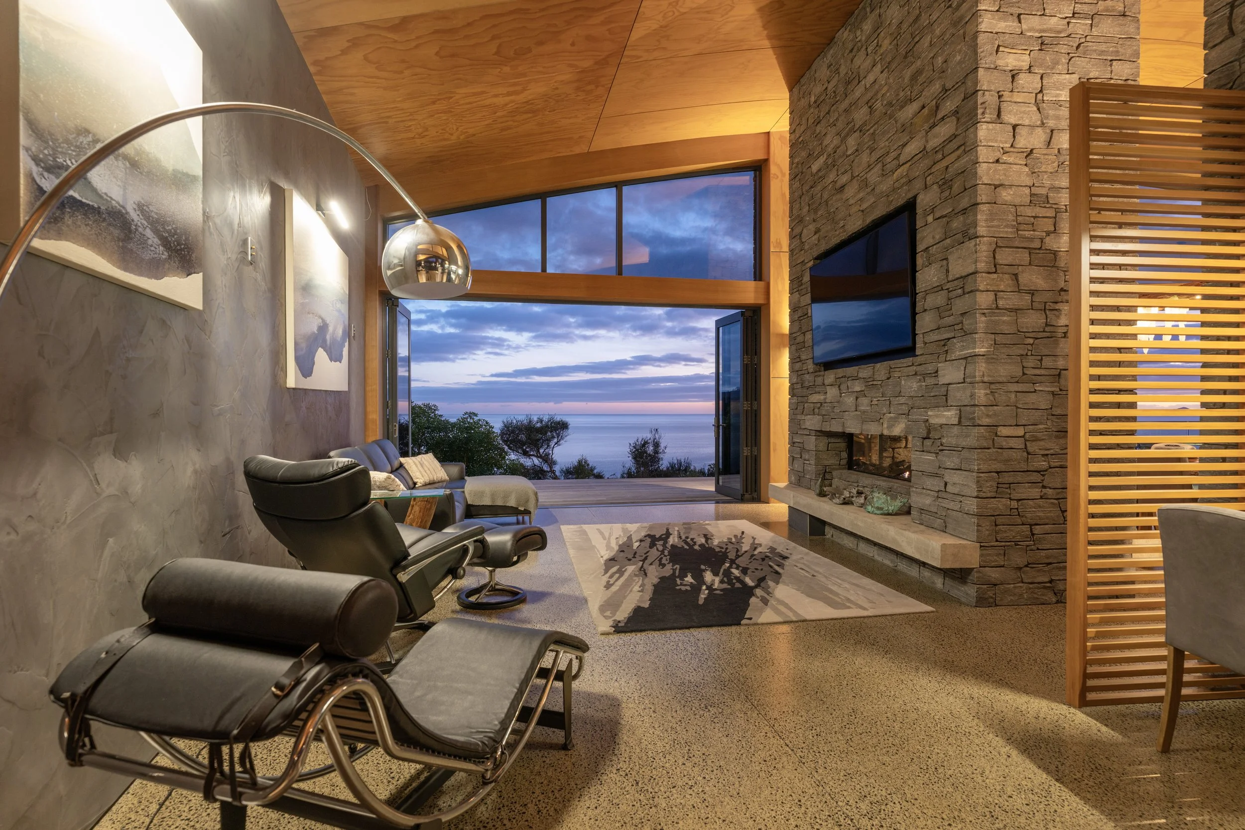 Modern living room with a stone fireplace, a large window with a view of the ocean at sunset, and contemporary furniture including a black lounge chair and a patterned rug.