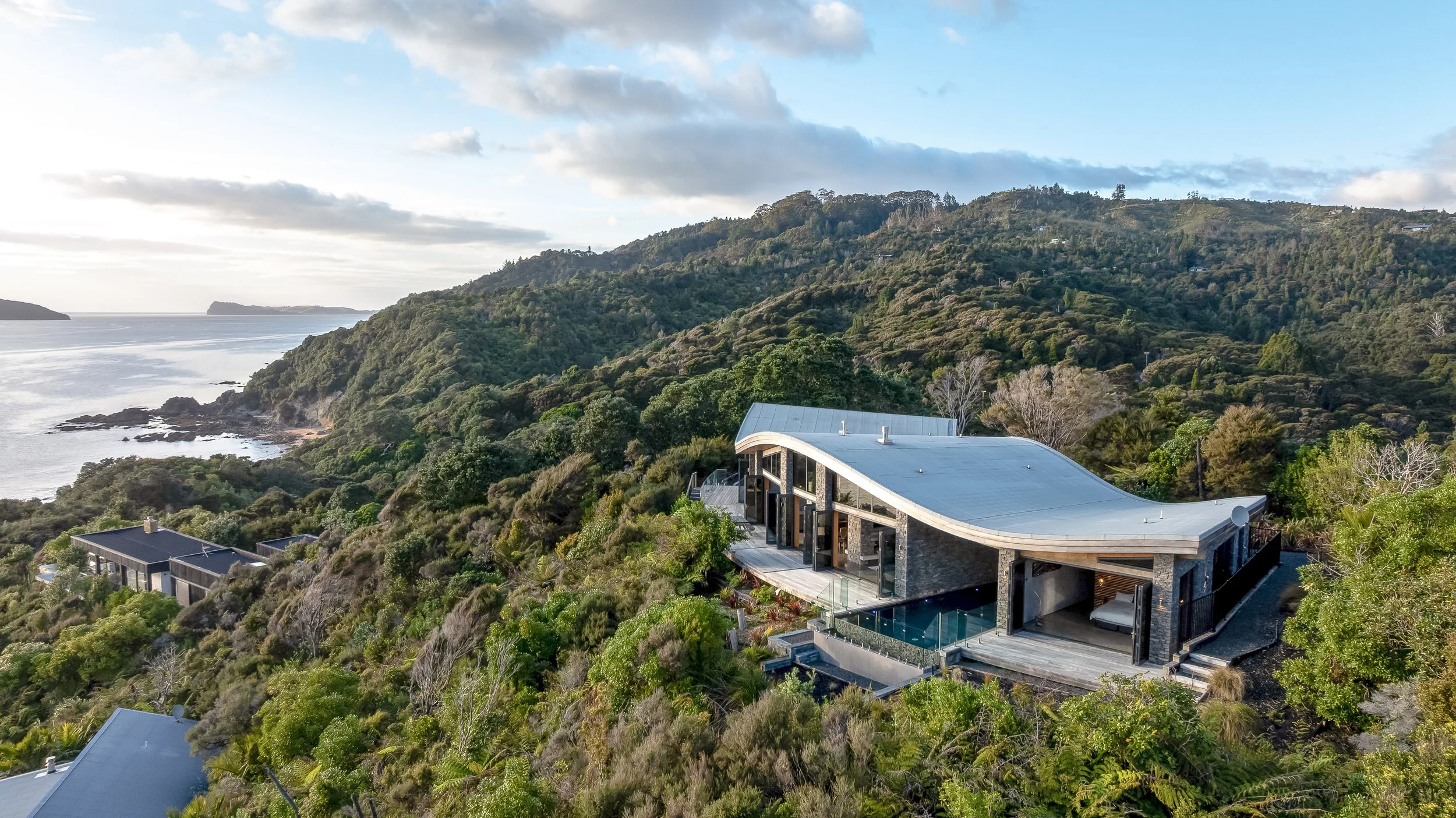 Modern house with a curved roof, large windows, and a swimming pool on a hill overlooking the ocean and lush green mountains.