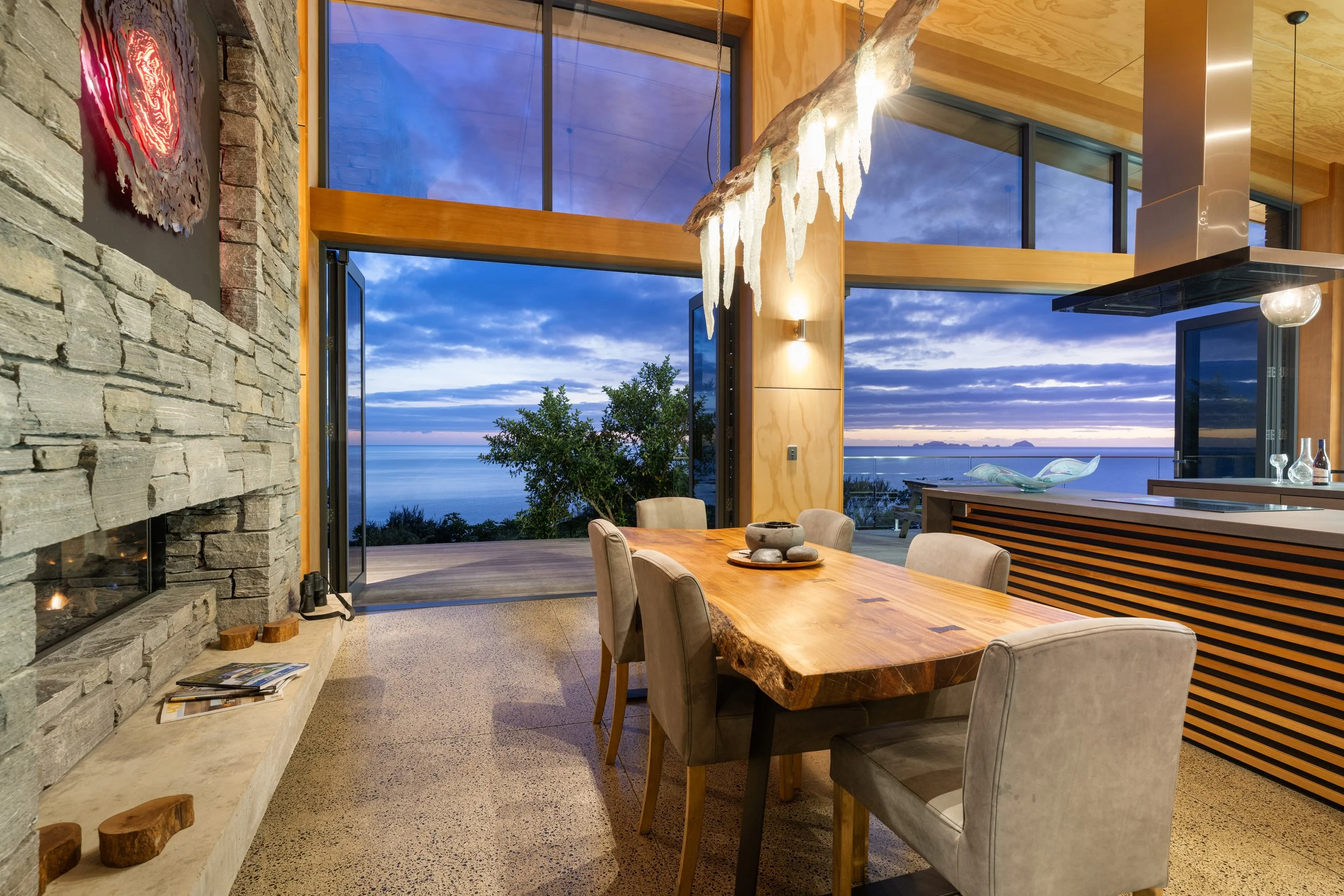 Interior of a modern dining area with a wooden table, beige chairs, stone fireplace, and large windows showcasing a scenic ocean view at sunset.