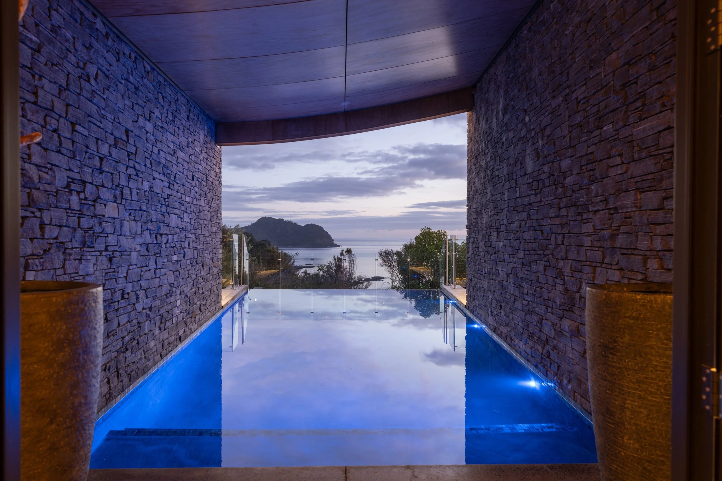 View of a private infinity pool overlooking a scenic coastal landscape with hills, trees, and water during dusk.