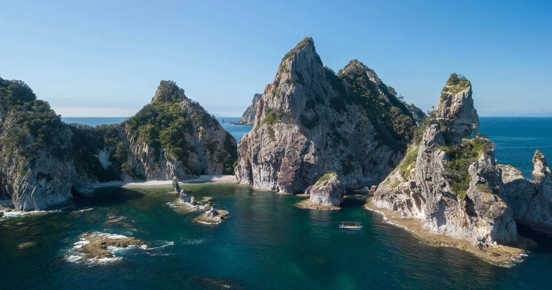 Aerial view of jagged rocky islands with green vegetation surrounded by blue water, with a boat near the shoreline and ocean in the background.