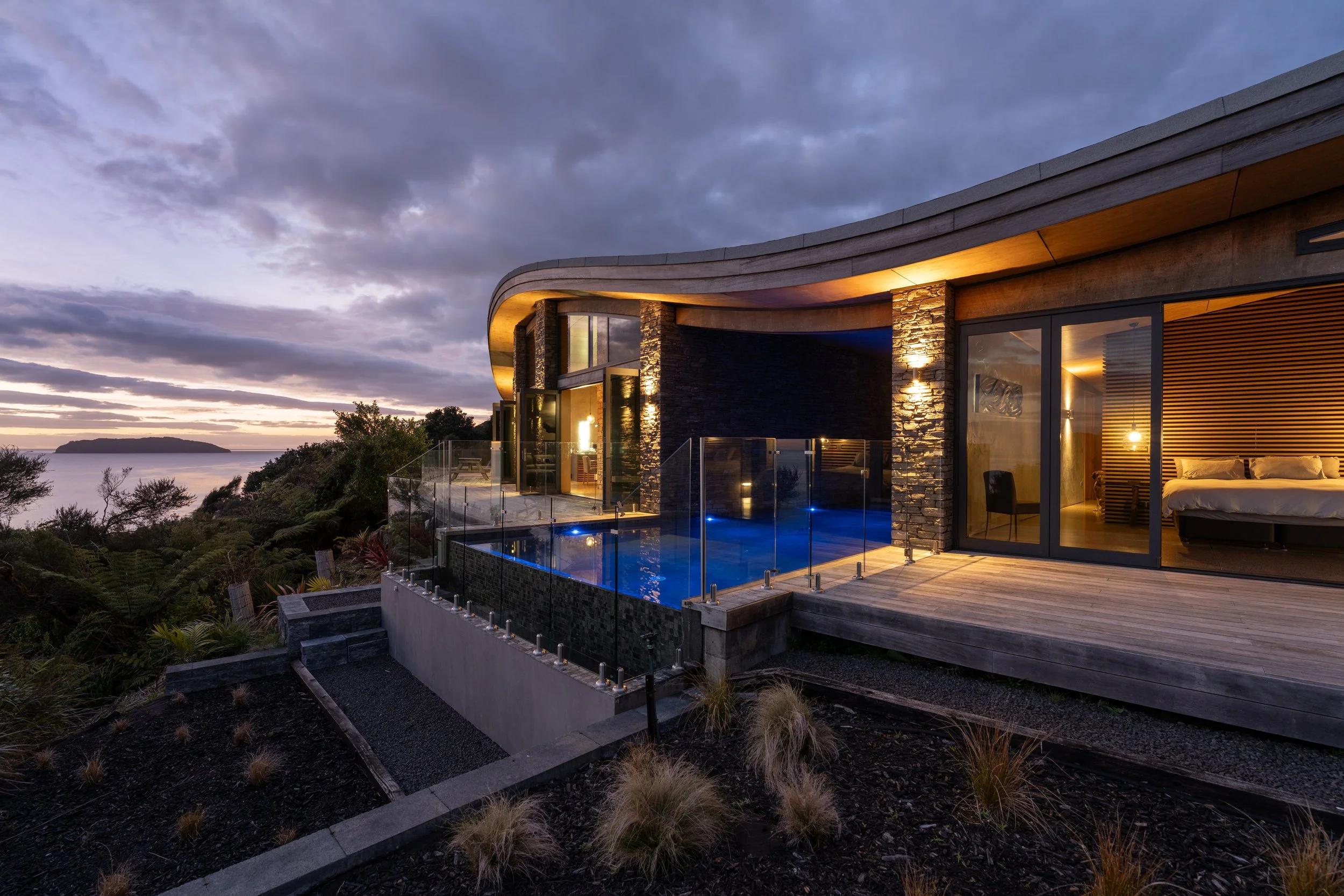 Modern house on a hillside during sunset with a glass balcony, outdoor pool, and large interior window showing a bedroom.