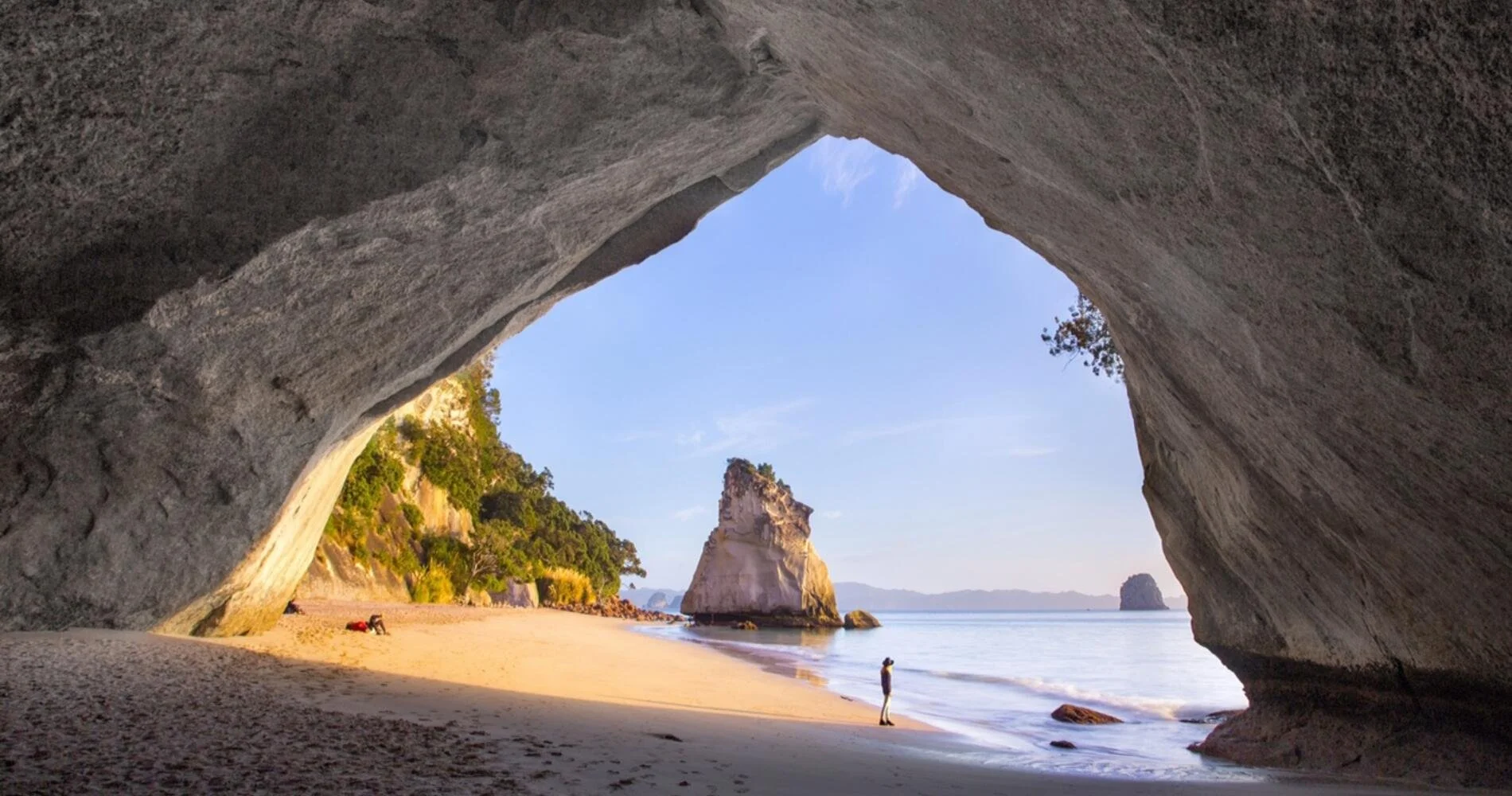 View through a large rock arch on a beach with a person standing at the shoreline, and a large rock formation in the water, with trees and a hillside in the background.