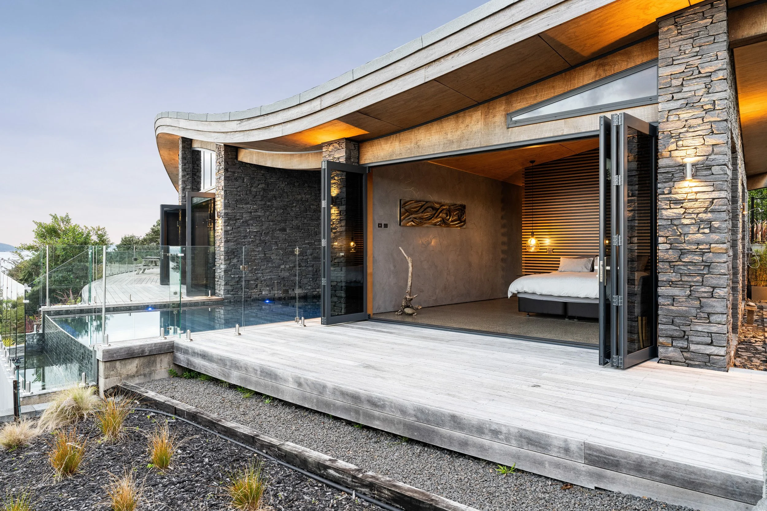 Modern house with a curved roof, stone and wood exterior, open sliding glass door revealing a bedroom with wooden wall accents and a bed, outdoor deck with glass railing, pool, and landscaped garden.