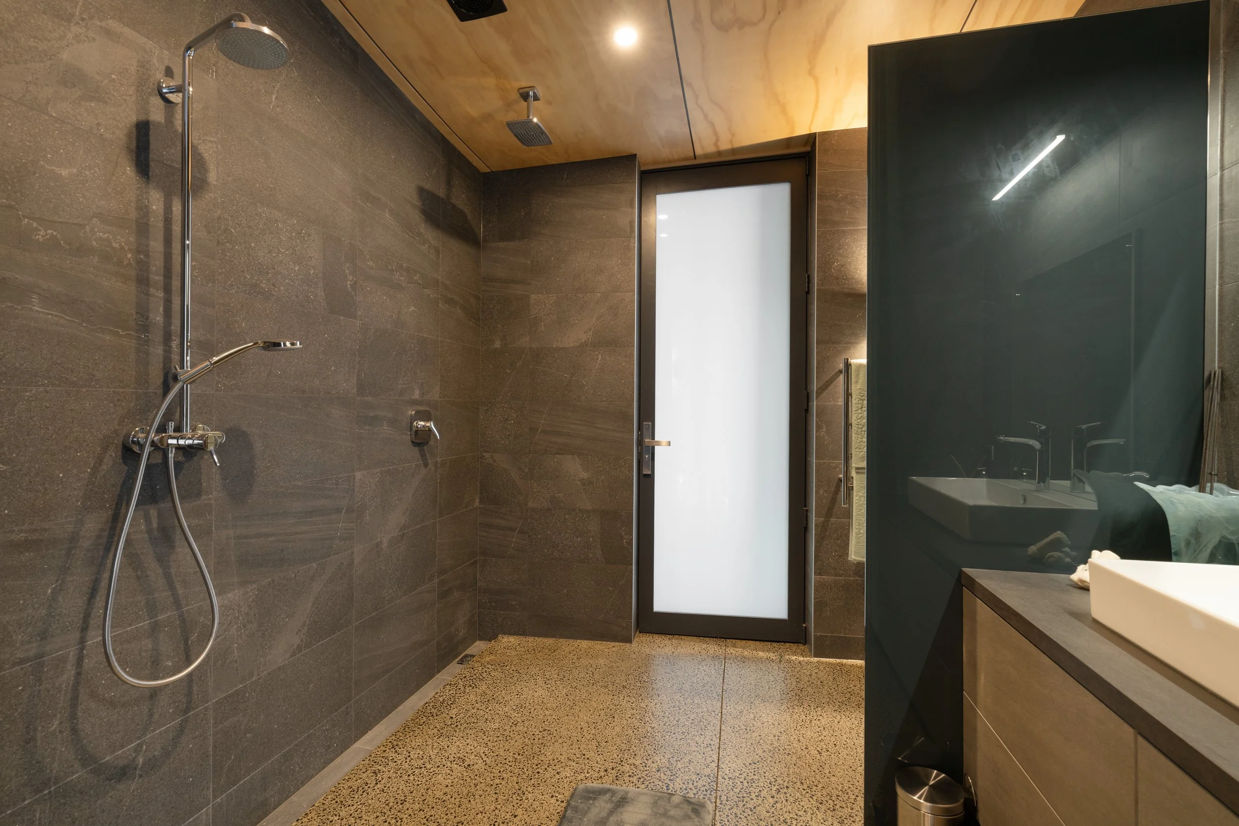 Modern bathroom with a walk-in shower featuring dark gray tiles, a handheld showerhead, and a ceiling rain shower. There is a frosted glass door leading outside or to another room. To the right, a vanity with a rectangular sink, a dark cabinet, and a mirror reflected in a glossy dark panel.