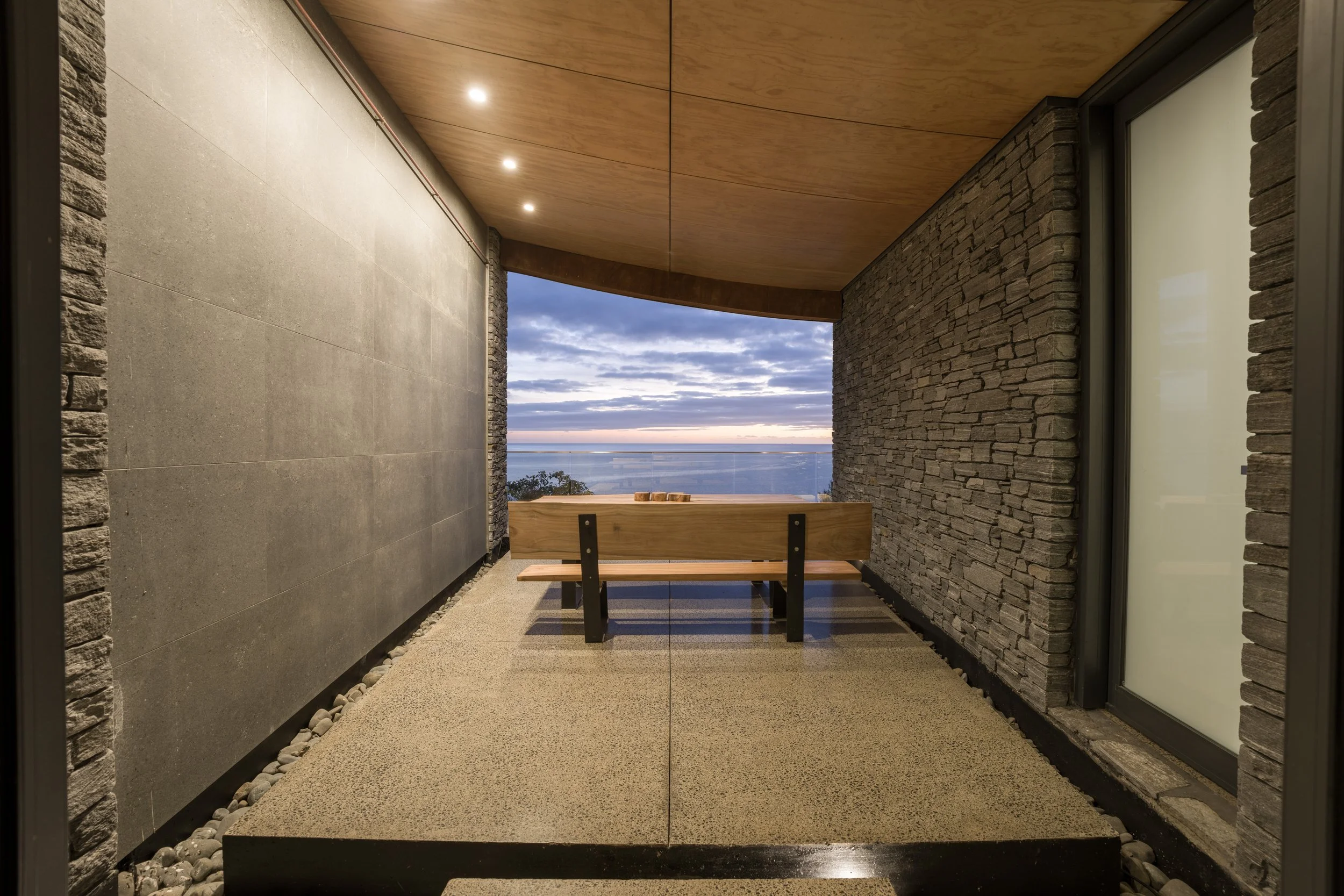 Balcony with a wooden bench overlooking a body of water and a sky with clouds at sunset.