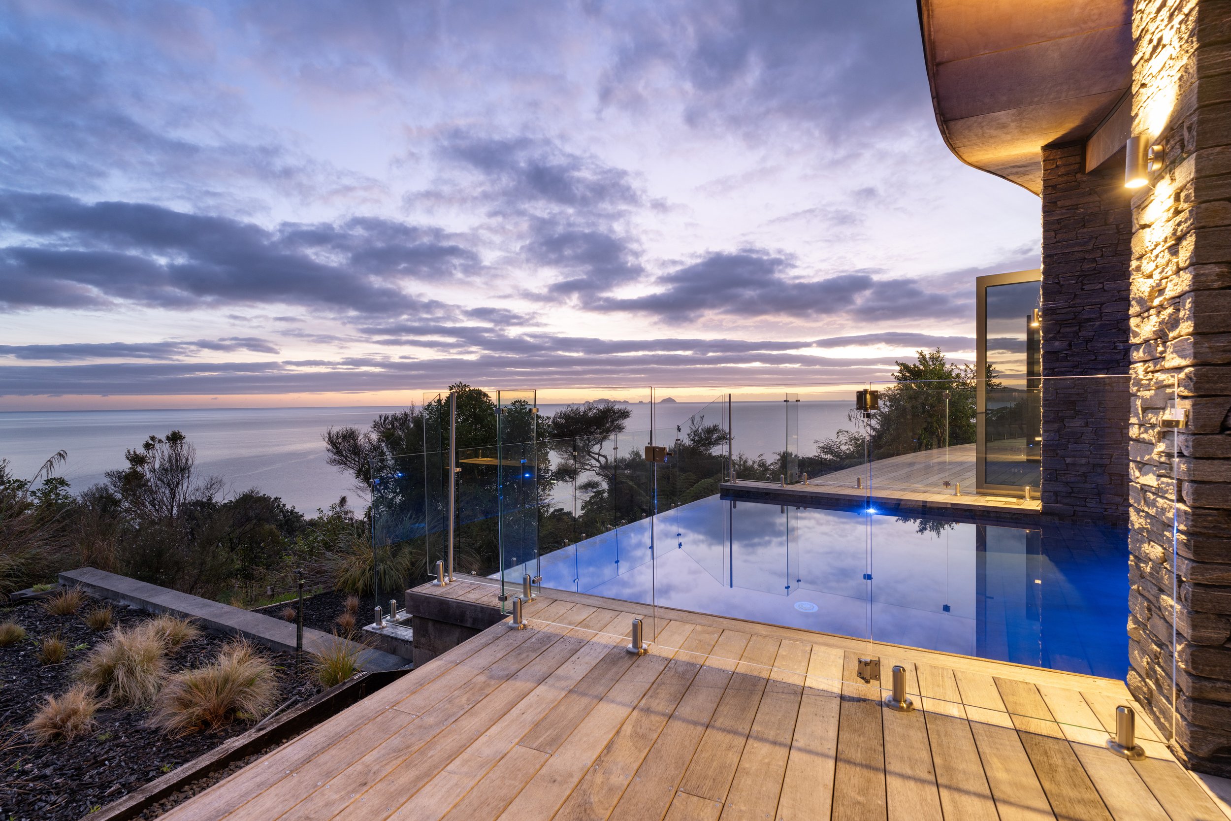 Scenic view of a modern infinity pool on a wooden deck, overlooking the ocean during sunset, with trees and cloudy sky in the background, and part of a house with stone wall and glass door.