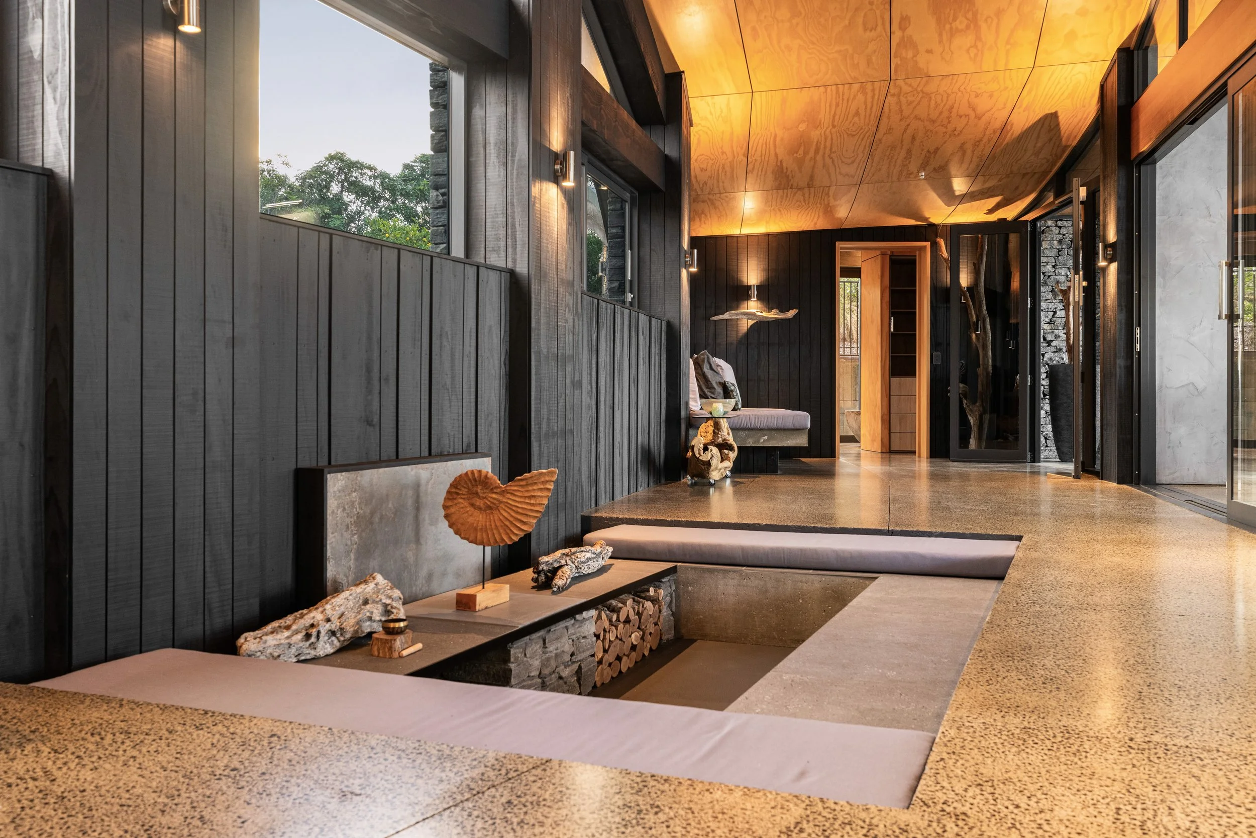 Modern interior living space with black wood panel walls, large windows, a built-in seating area, decorative art pieces, and warm lighting.