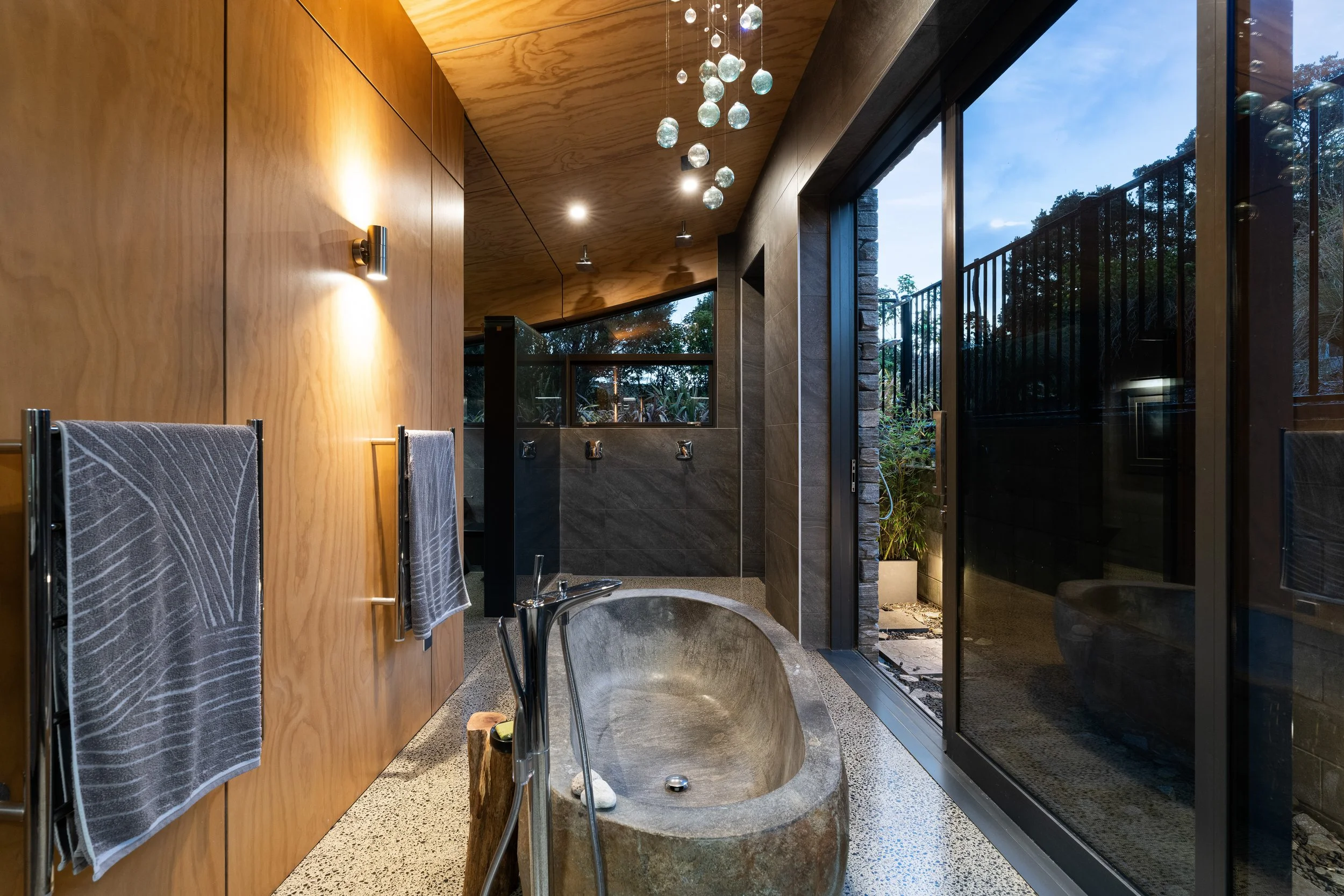 Modern bathroom with a freestanding stone bathtub, wooden ceiling and wall panels, and large sliding glass door showing a patio and garden outside. Towel racks with towels and hanging light fixtures are visible.