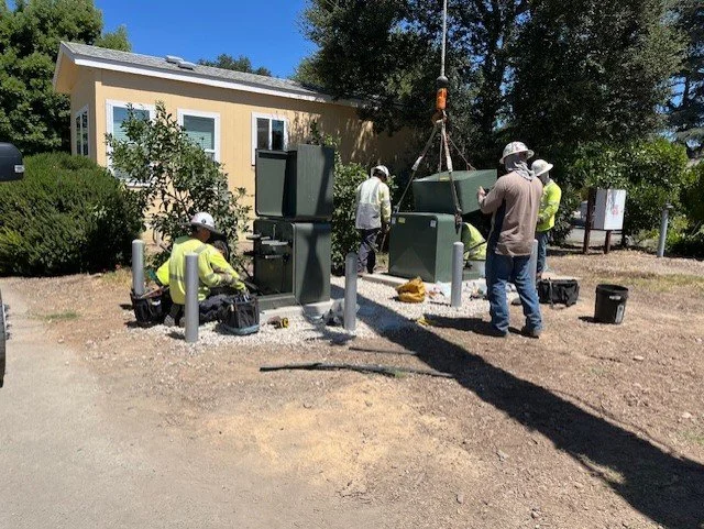 Workers wearing safety helmets and reflective vests installing or repairing electrical or communication equipment outdoors near a small building.