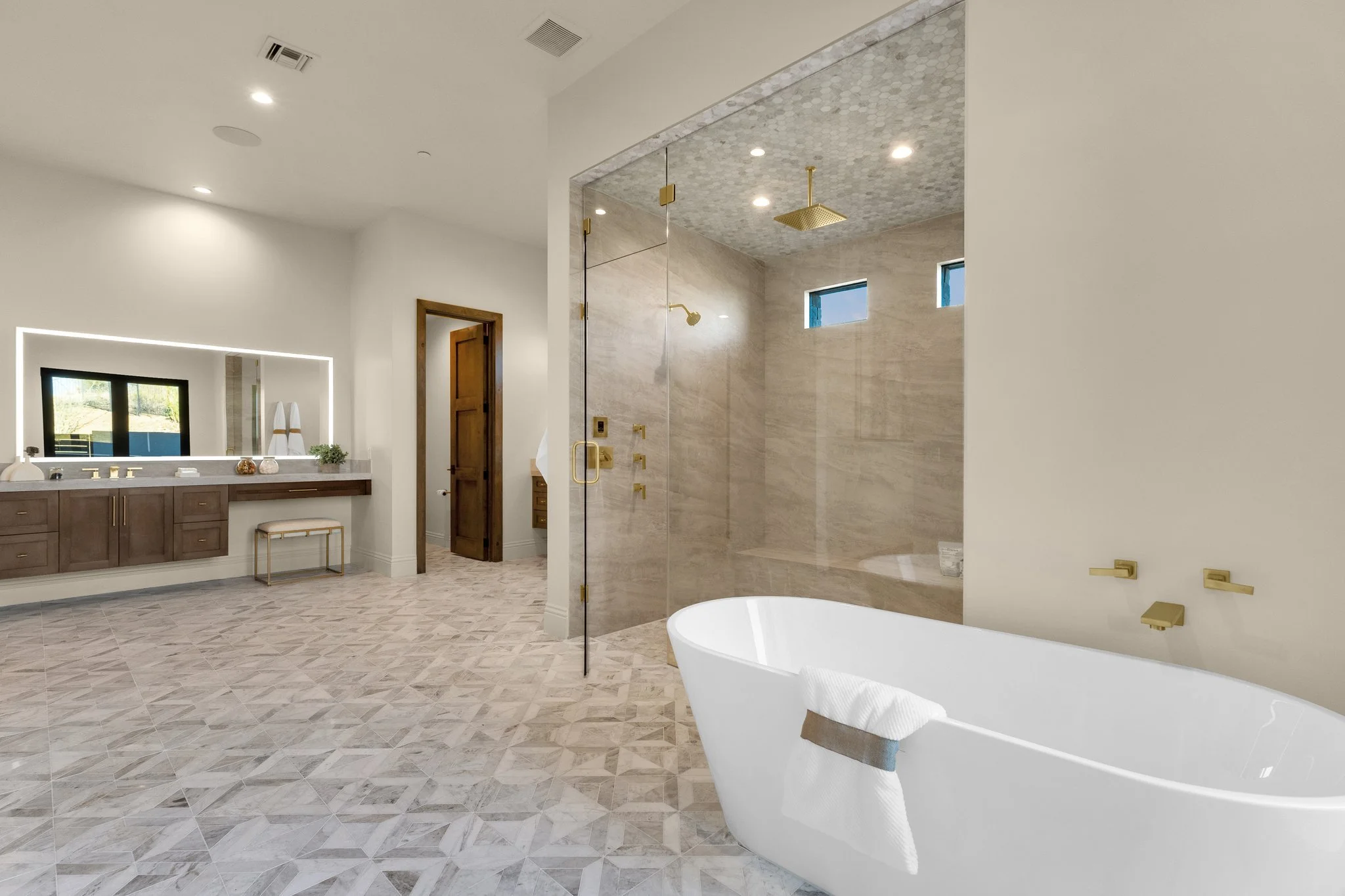 Modern bathroom with a white bathtub, geometric tiled shower area with brass fixtures, custom wooden vanity with a large LED mirrors.