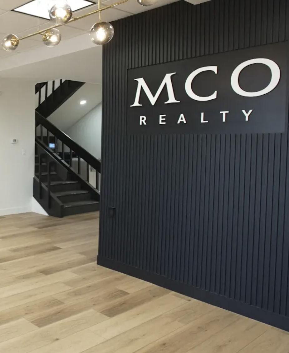 Interior of a real estate office with a black accent wall, wood flooring, and a staircase leading to an upper level. The wall features a sign reading 'MCO Realty' in large white letters. Modern ceiling light fixtures are also visible.