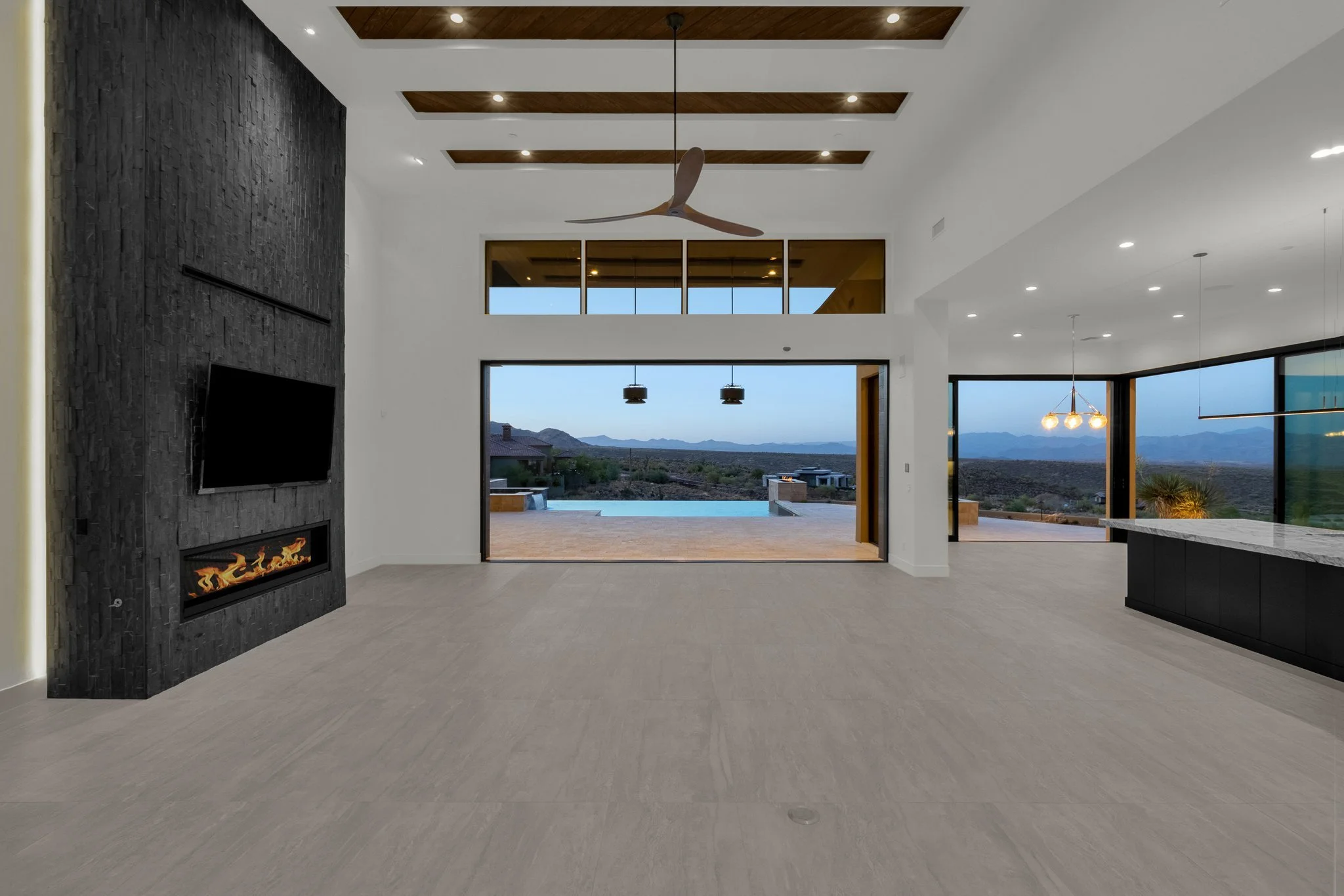 Modern living room with large windows overlooking desert landscape, featuring a black stone fireplace, flat-screen TV, ceiling fan, and open floor plan.
