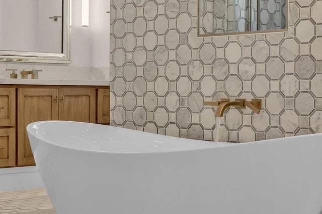 A bathroom with a freestanding white bathtub, wood vanity with a white countertop, and a wall covered with marble patterned tiles. There is a brass wall-mounted faucet and a mirror above the vanity.
