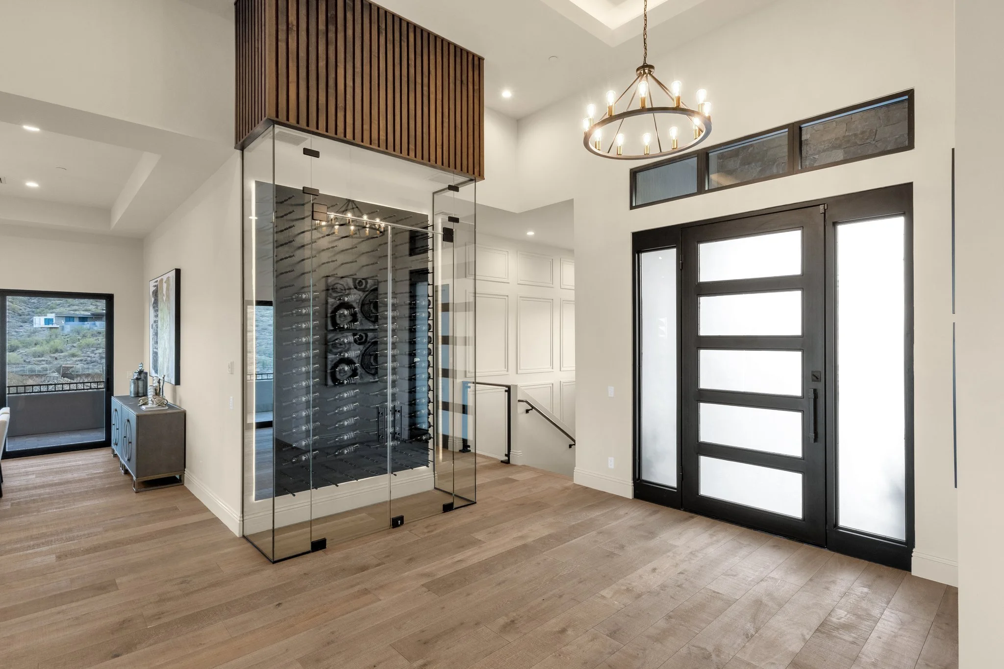 Modern entryway with black front door, frosted glass panels, and a chandelier. Wine cellar with glass walls and wine bottles inside. Light wood flooring, staircase, and decorative paneling.