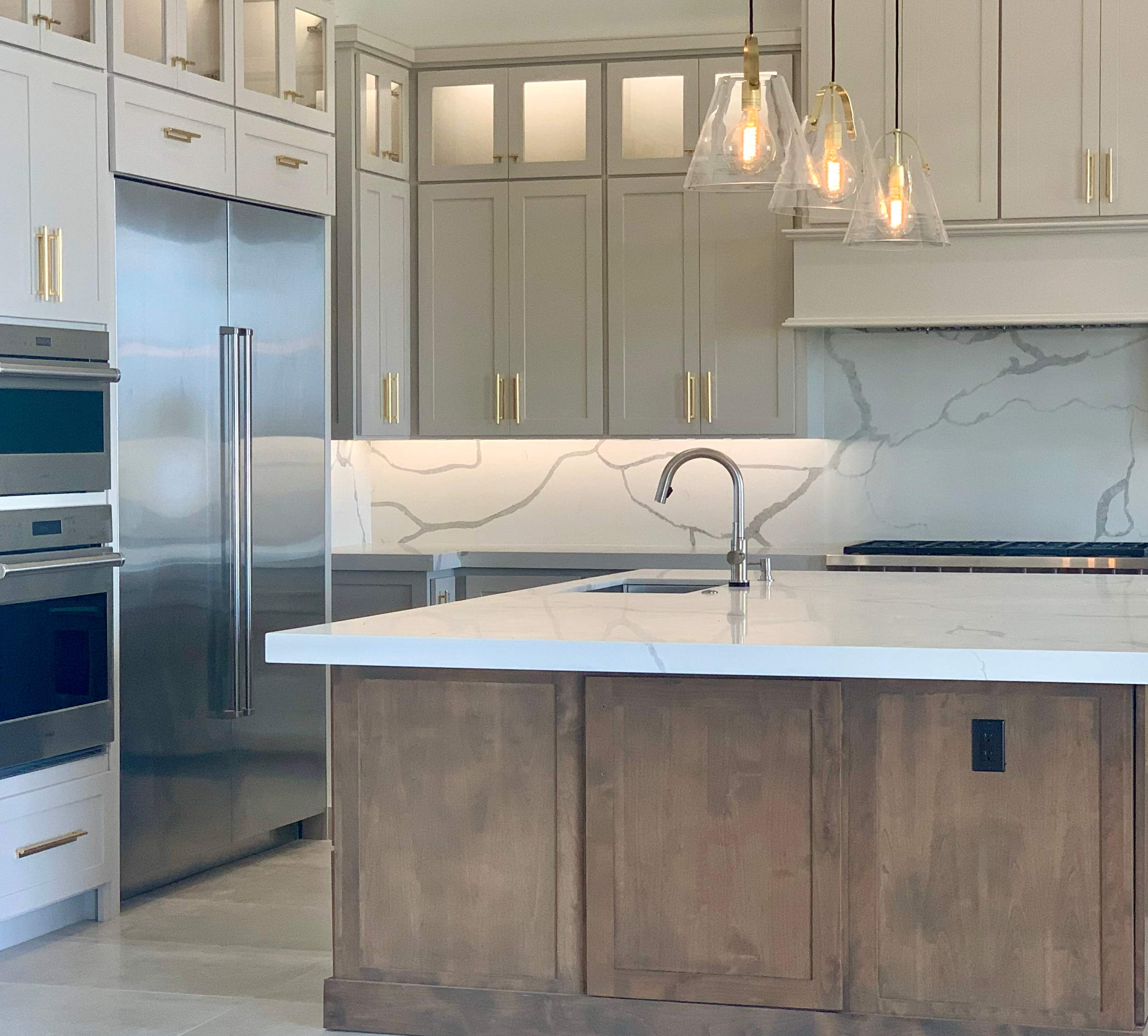 Modern kitchen with greige cabinets, a white quartz backsplash, a white countertop, a wooden island with a white countertop, and stainless steel appliances, including a double oven and a refrigerator. 