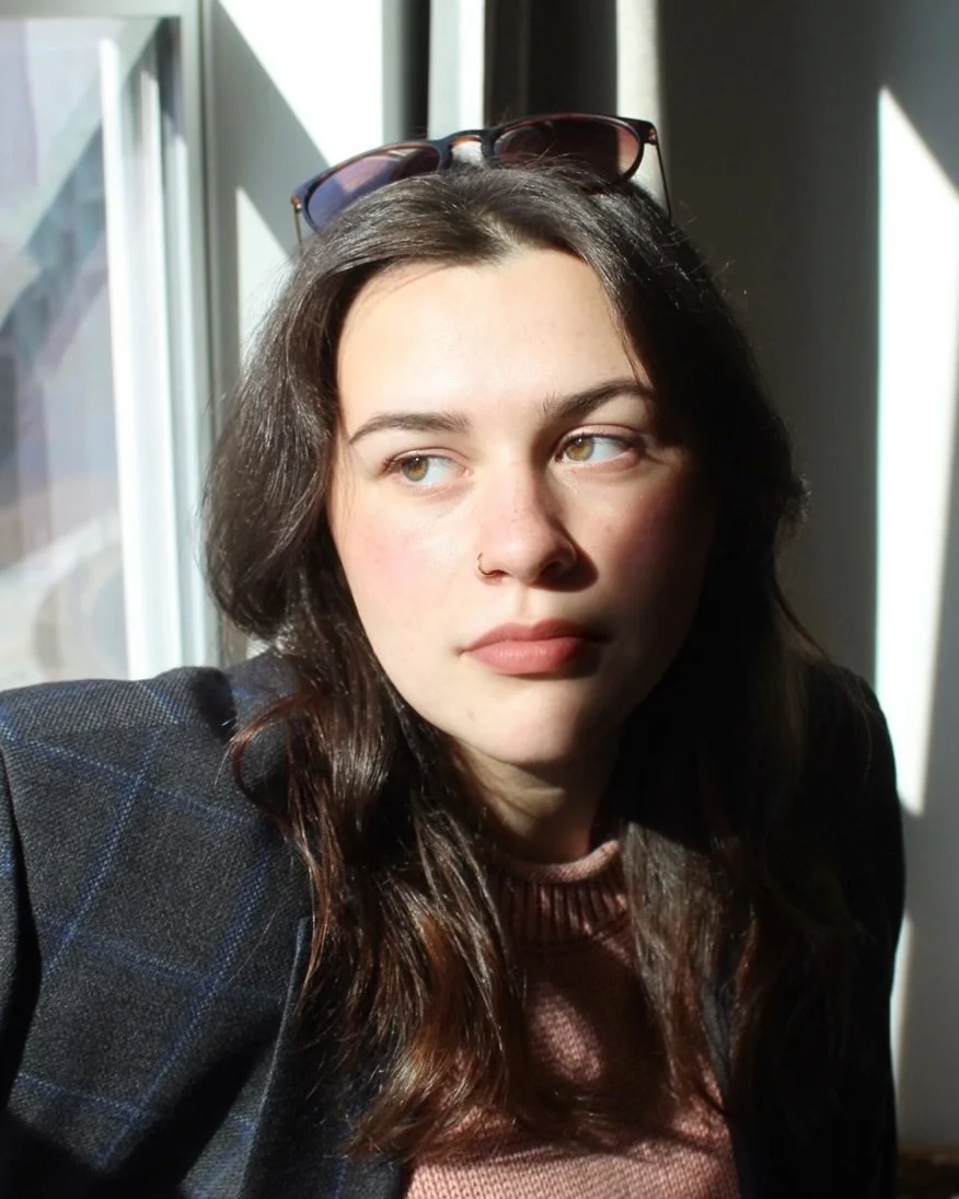 A young woman with long dark hair, light skin, and hazel eyes, sitting by a window with sunlight on her face, wearing sunglasses on her head, a nose ring, a dark blazer, and a mauve top.