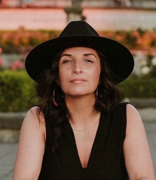 A female Canadian Financial Broker with wavy dark hair, wearing a black sleeveless top, large hoop earrings, a delicate necklace, and a wide-brimmed black hat, sitting outdoors with a blurred background of flowers and a water feature.