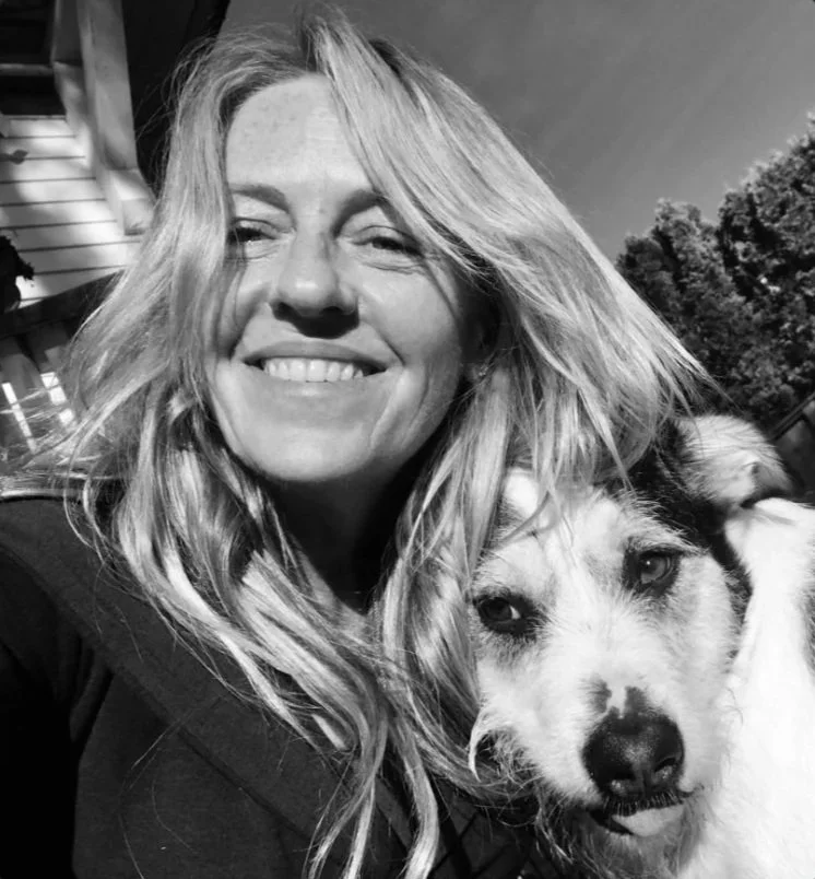 A female Canadian Financial Broker with long wavy hair smiling and taking a selfie with her dog outdoors, with trees in the background.