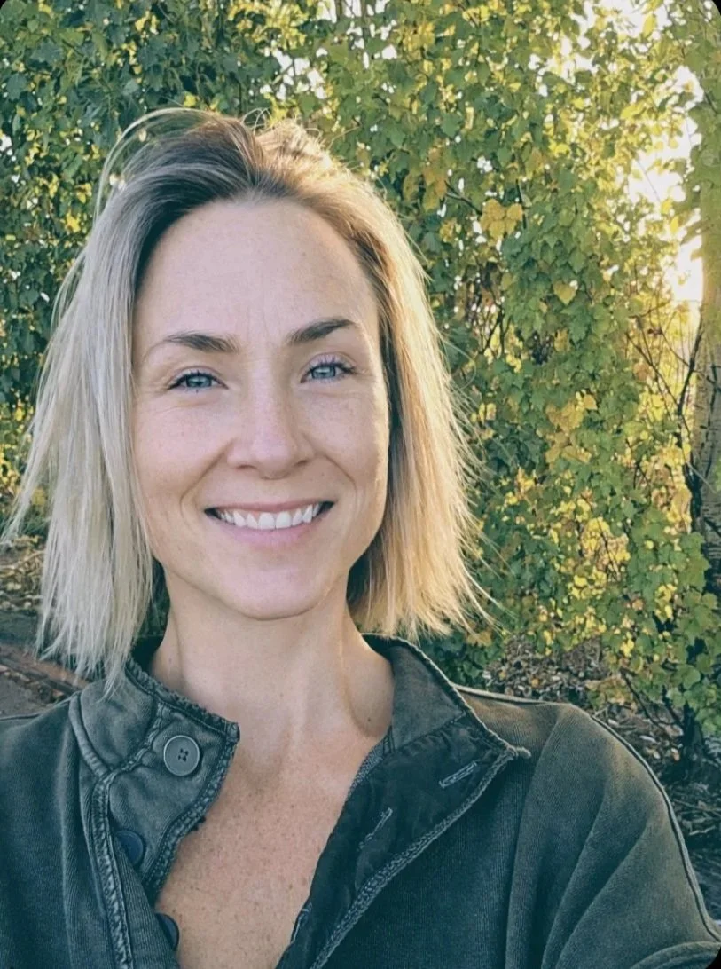 A female Canadian Financial Broker with blonde hair smiling outdoors in front of green foliage.