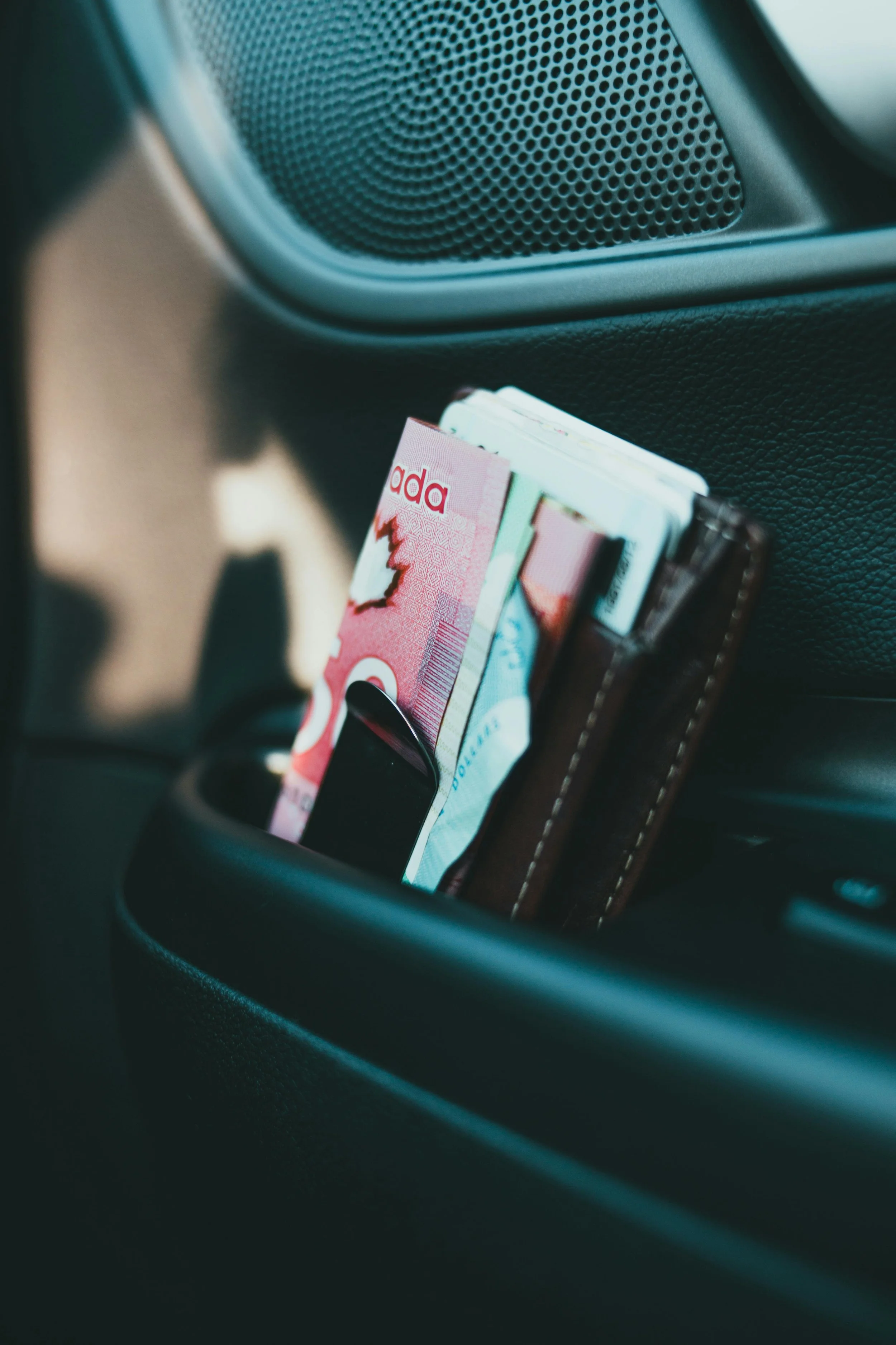Car door pocket holding paper currency, a smartphone, and a wallet, with a speaker grille visible above.