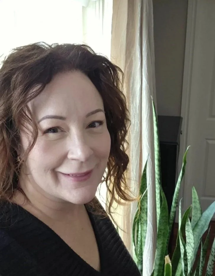 A female Canadian Financial Broker with curly brown hair smiling, standing indoors near a window with cream-colored curtains and a houseplant with long green leaves.
