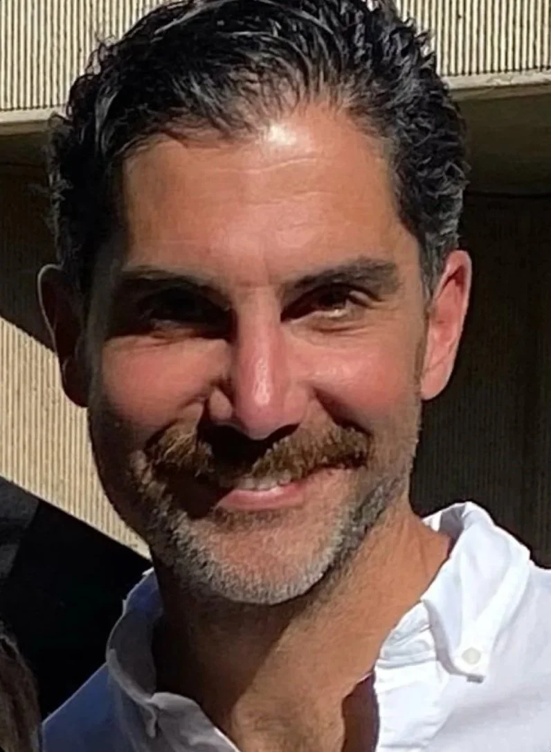 A smiling middle-aged man Canadian Financial Broker with dark, wavy hair, a beard, wearing a white shirt, outdoors with a building in the background.