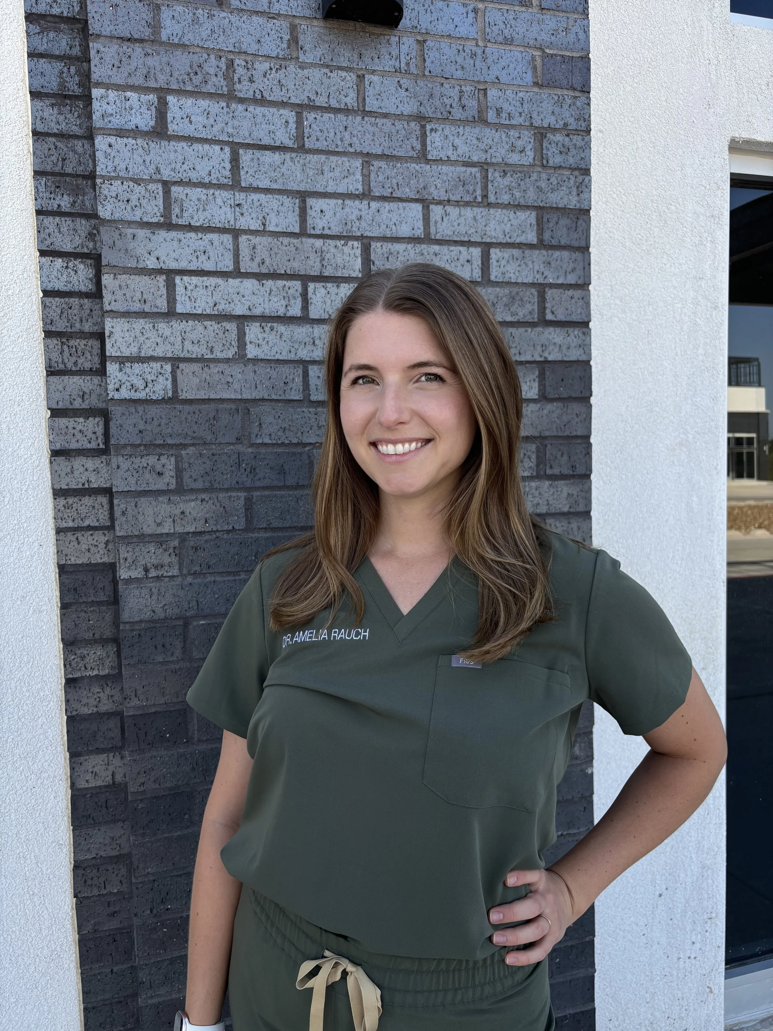 This is a photo of Amelia Rauch, a veterinarian in Boerne, Texas posing in front of a dark brick wall and wearing green scrubs.