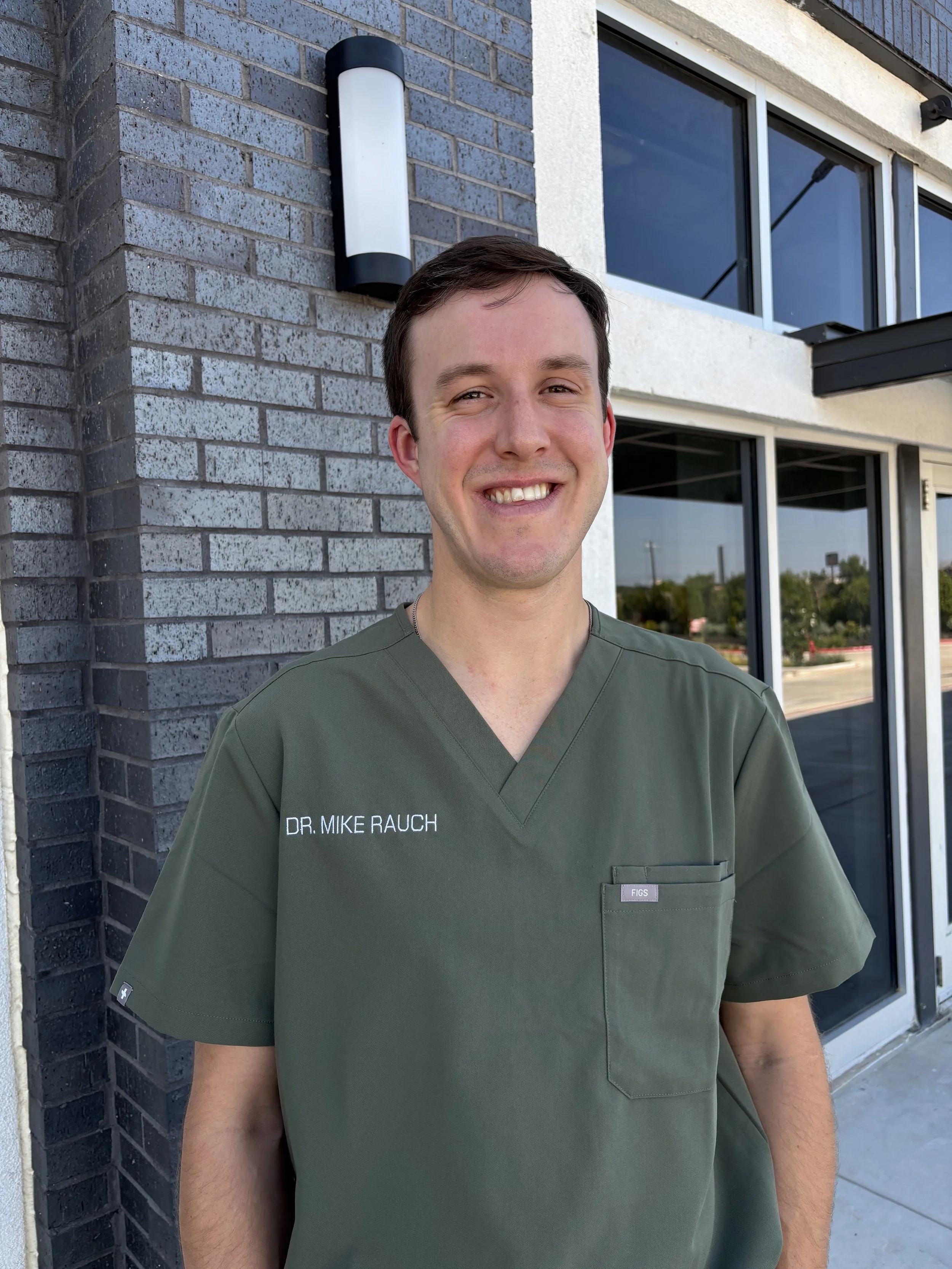 This is a photo of Mike Rauch, a veterinarian in Boerne, Texas, posing in front of glass windows and wearing green scrubs.