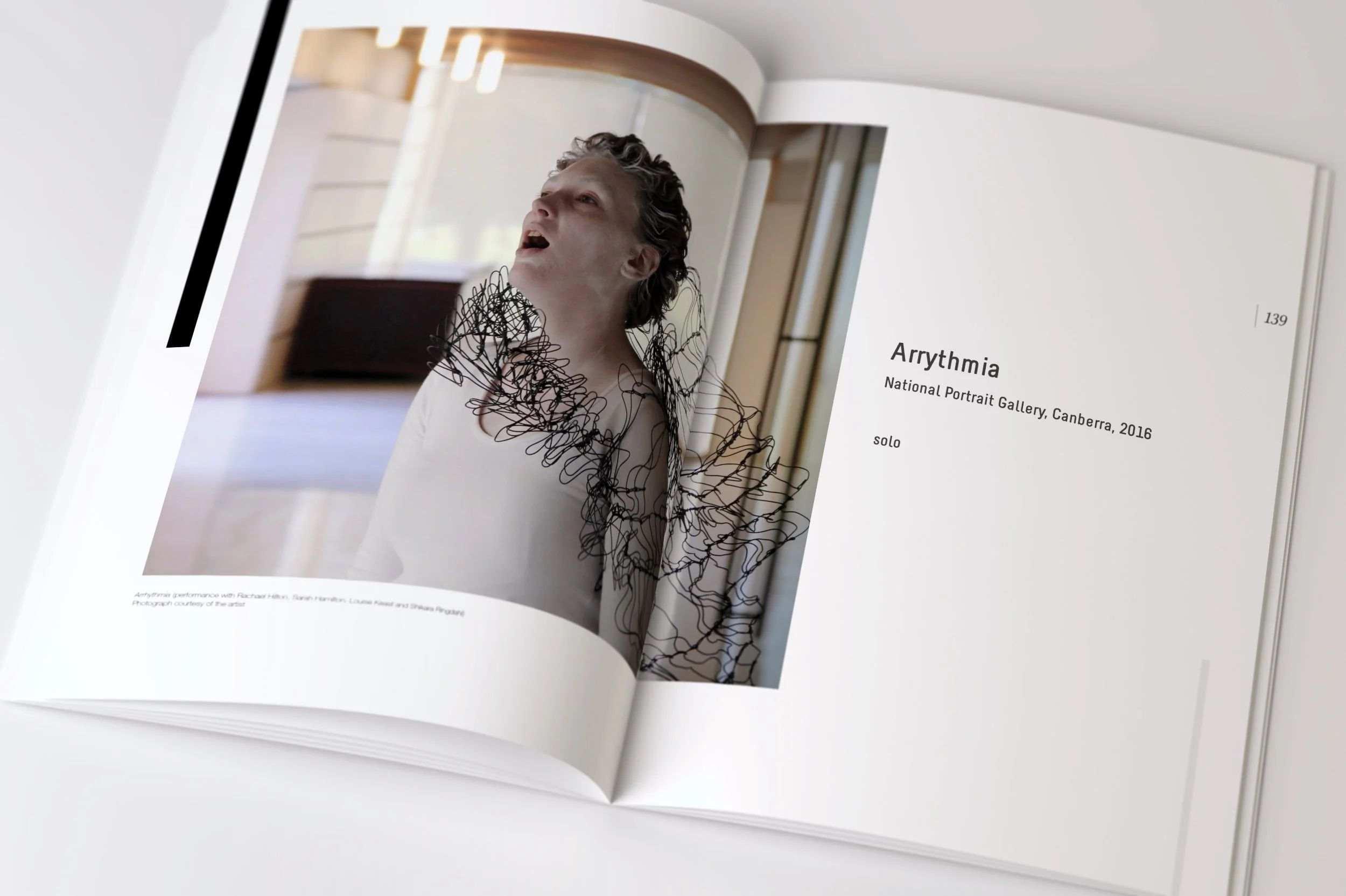 A photo of an open art book showing a photograph of a performer with styled hair, wearing a shirt with a black wire-like art piece around their neck and shoulders, titled 'Arrythmia' from the National Portrait Gallery, Canberra, 2016.