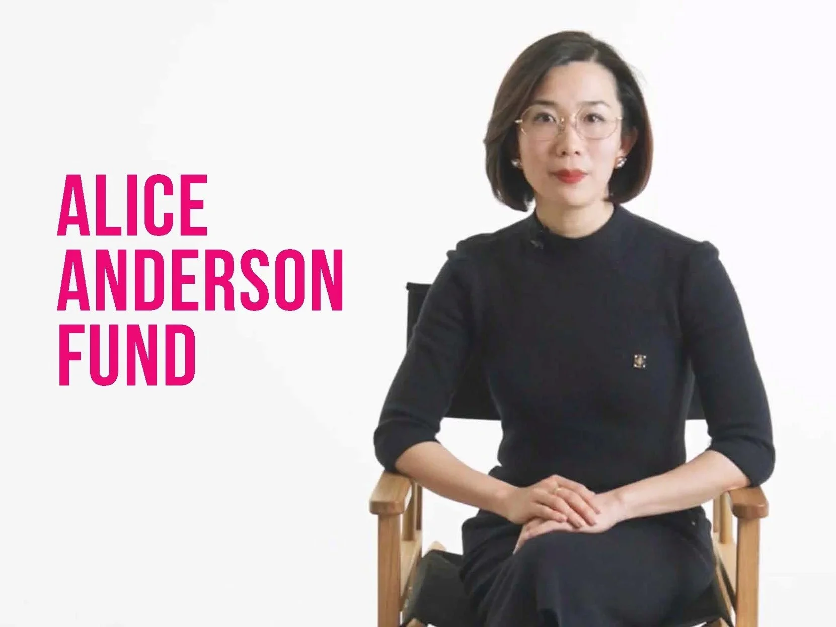A woman with glasses and shoulder-length dark hair sitting on a wooden chair against a white background. She is wearing a black dress with three-quarter sleeves and has a serious expression. To her left, there is text in bold pink letters that reads 