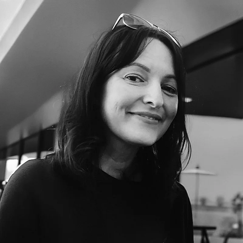 A woman with shoulder-length dark hair, wearing glasses on her head, smiling at the camera, in a black and white photo.