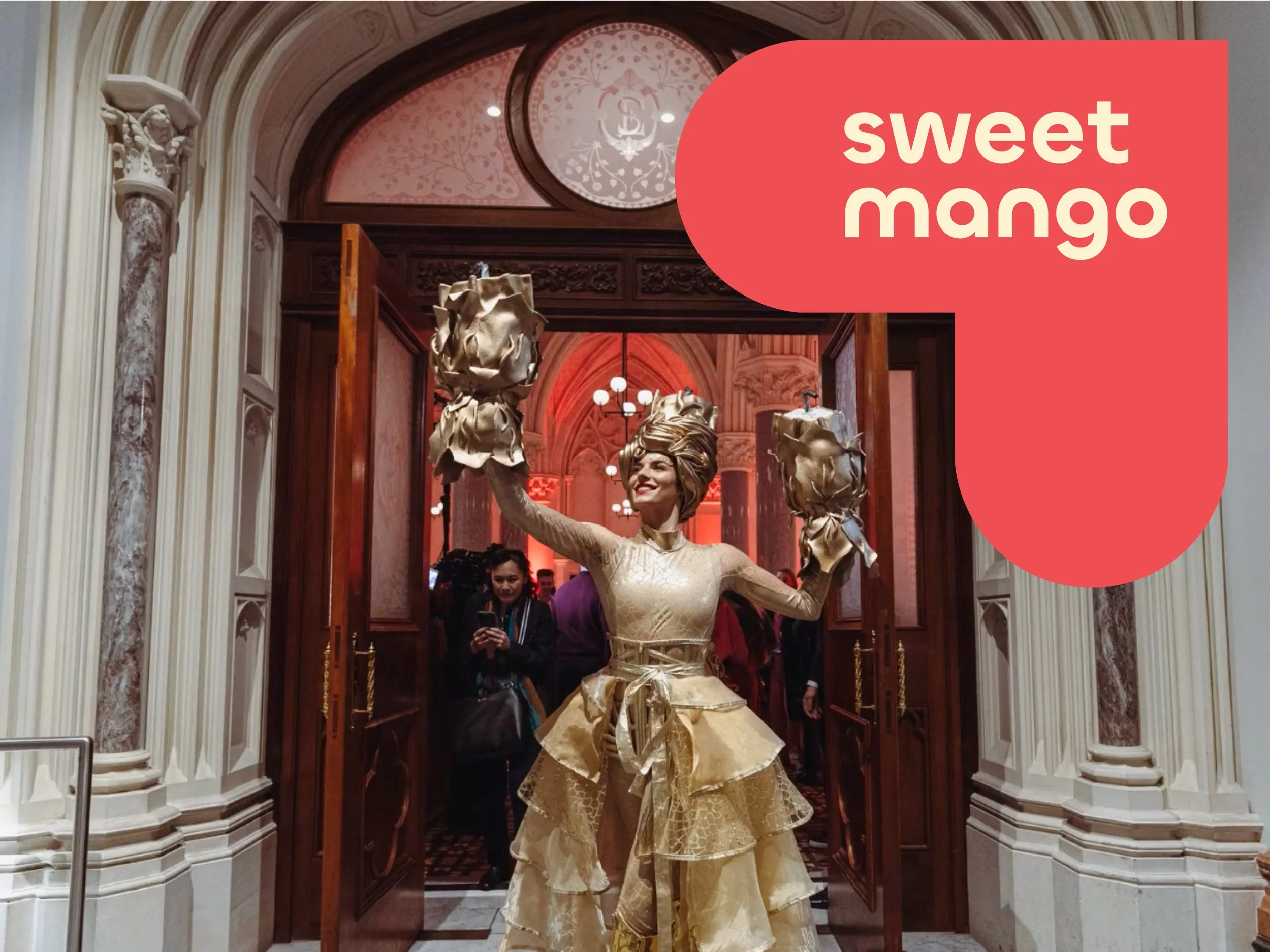 A woman in a gold and beige dress with large bows on her head poses in a grand, ornate hallway with wood accents and a high, arched ceiling.