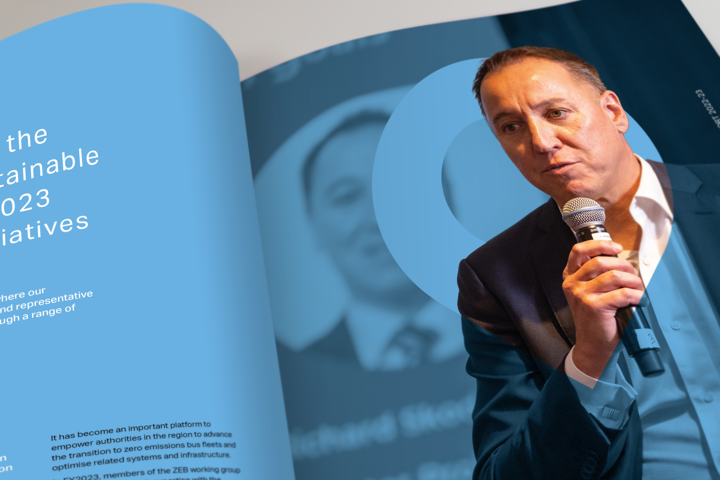A person in a suit holding a microphone and speaking at an event, with a blue and white branding backdrop in a publication.