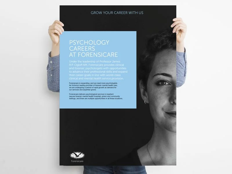 A person holding a black and blue poster that advertises psychology careers at Forensicare. The poster includes text about clinical and forensic psychology training and services, and features a half-face portrait of a woman on the right side.