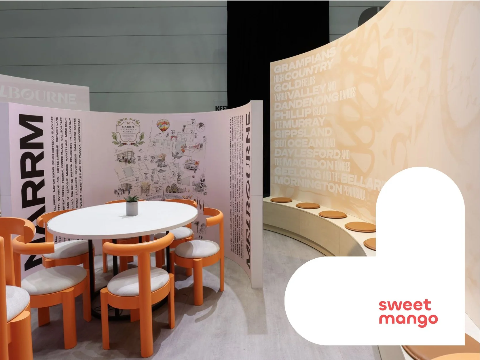An exhibition stand containing a modern interior space with a white round table and orange chairs, a curved seating area with orange cushions, decorative wall panels with text and graphics, and a small green potted plant on the table.
