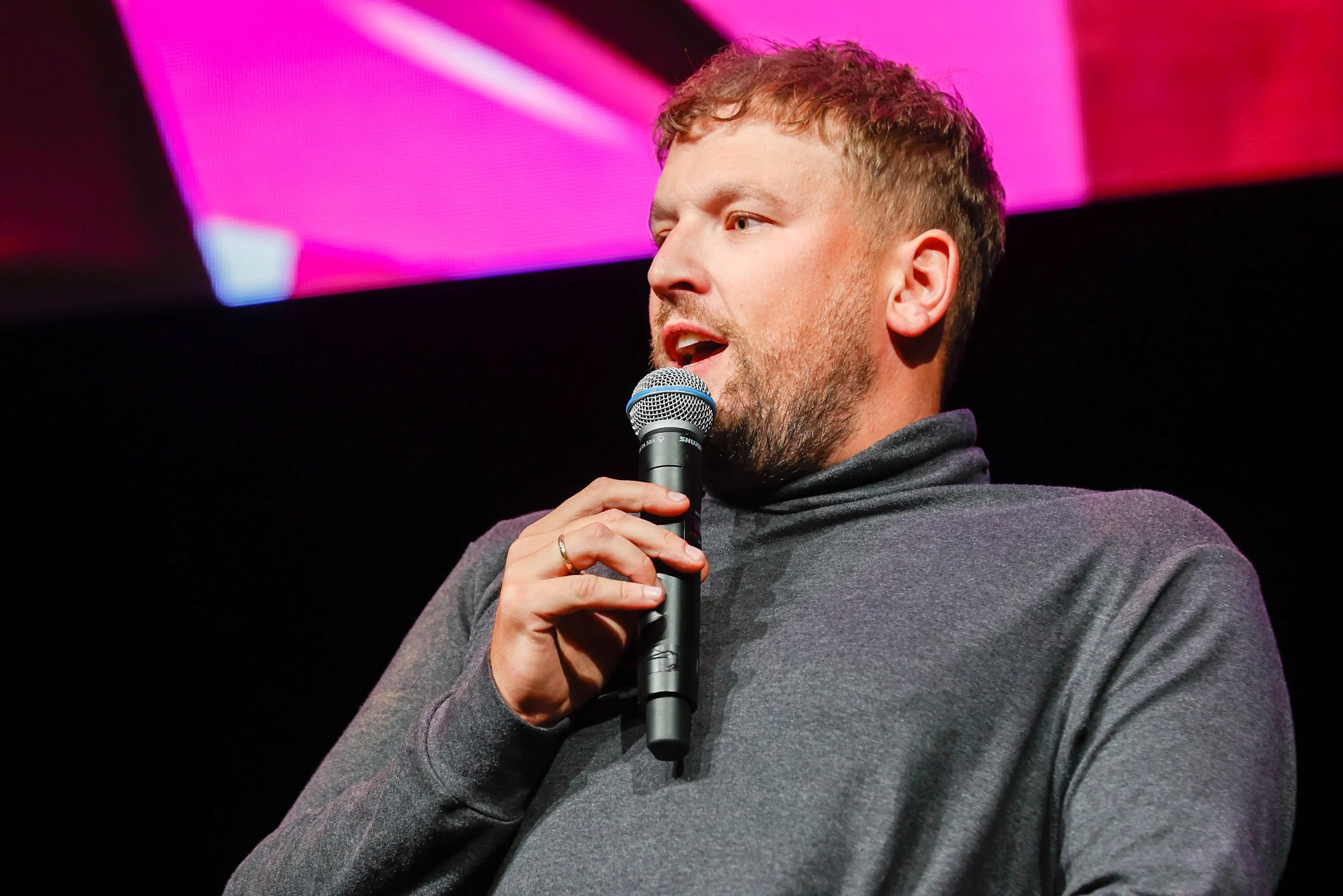 A young man with short curly hair and a beard speaking into a microphone on stage, with a background illuminated by pink and purple lights.