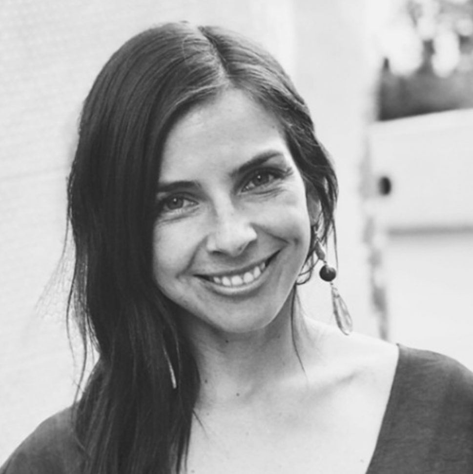 Black and white photo of a woman with long dark hair, smiling, wearing an earring, and a top with a wide neckline.