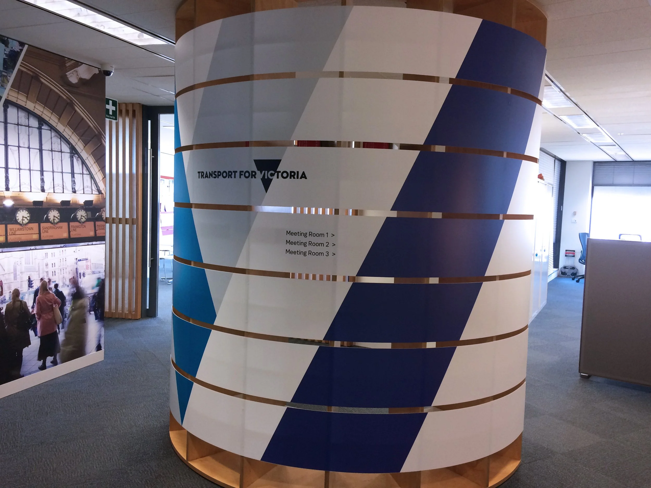 A cylindrical sign with blue, white, and beige design, displaying the Transport for Victoria logo and directions to meeting rooms in an indoor area of a transit or office building.