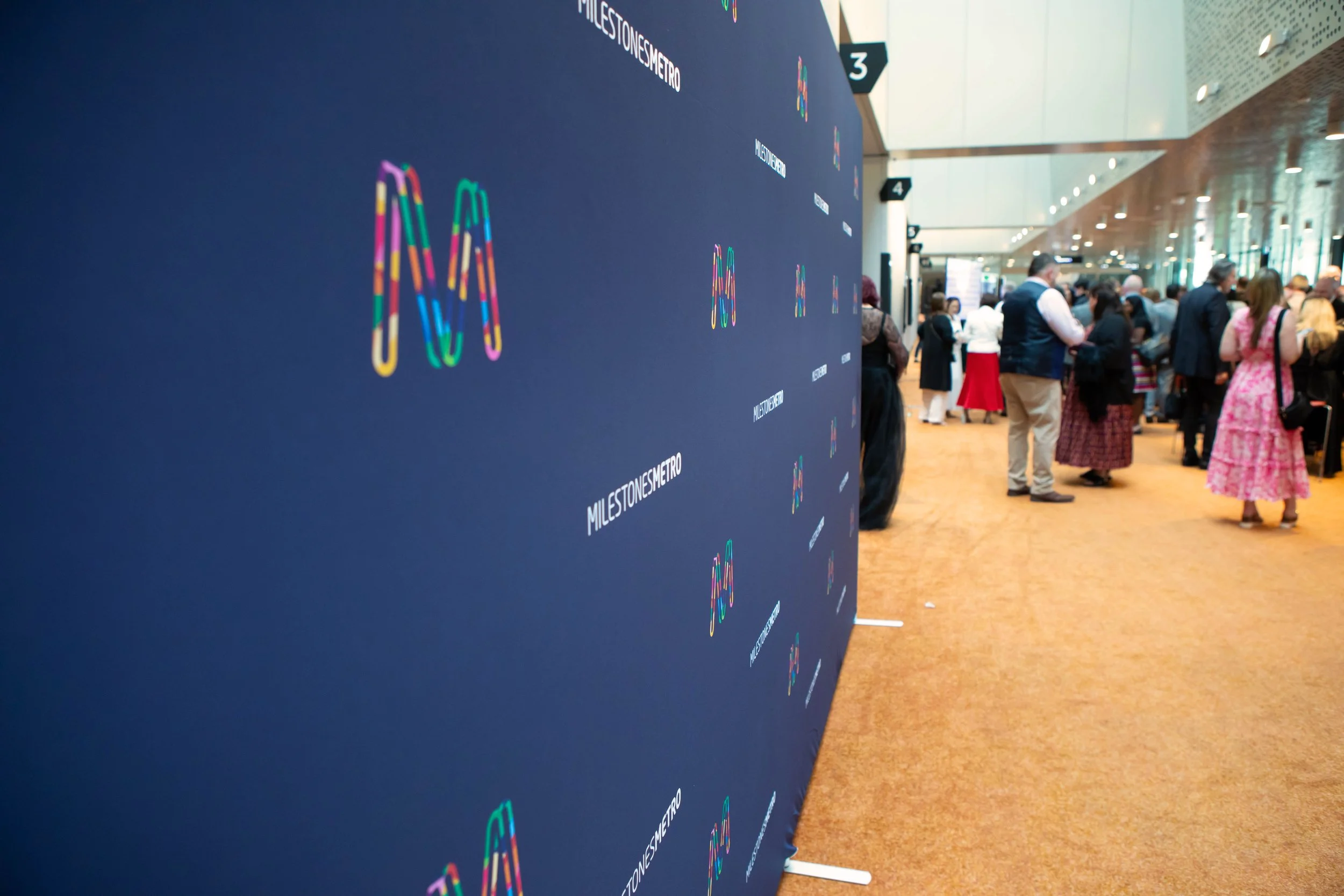 A large blue wall with colorful logos and text reading 'Milestones Metro' at an event or conference. In the background, a crowd of people are gathered in a spacious, well-lit hall.