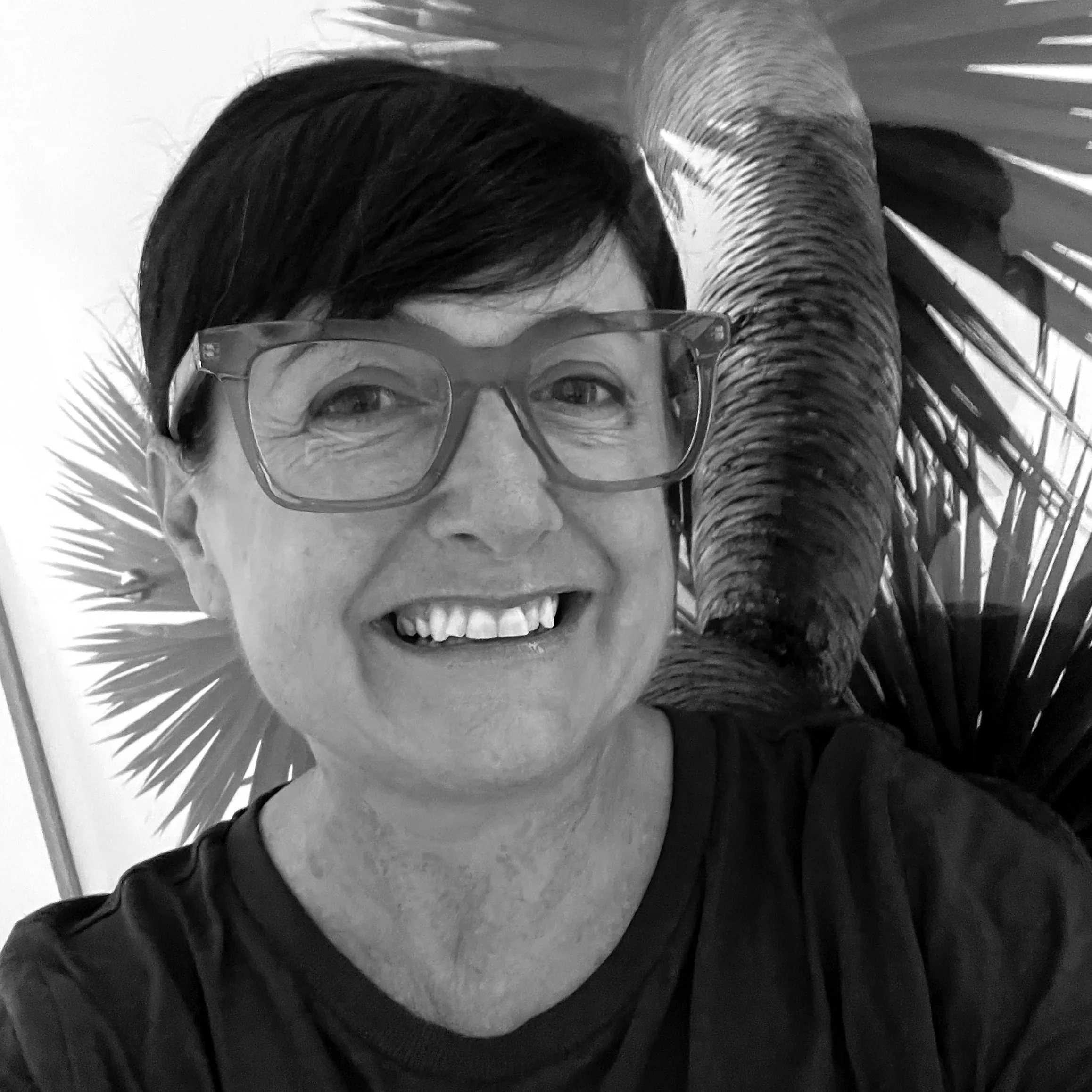 A woman with dark hair and glasses smiling behind a mask, with palm leaf decorations and a desk with some objects in the background.